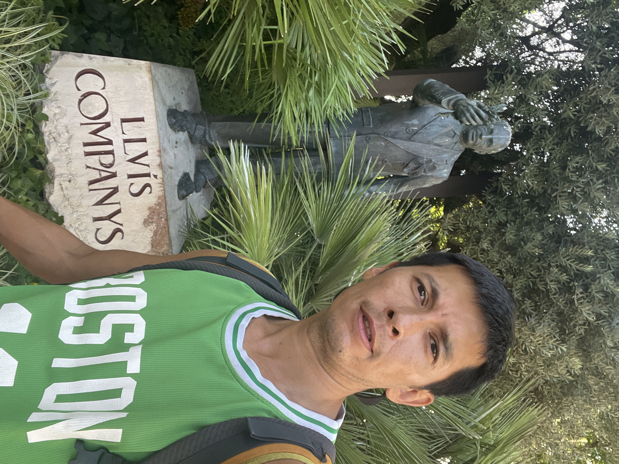 A man wearing a green Boston Celtics basketball jersey and a backpack poses for a selfie in front of a bronze statue of Lluís Companys. The statue stands on a stone base inscribed with "LLVIS COMPANYS". Lush green foliage, including palm fronds and trees, surrounds the statue and the man. The setting appears to be an outdoor memorial or garden, likely in Tarragona, Spain, given the presence of Lluís Companys, a significant figure in Catalan history who served as President of Catalonia. The man is looking directly at the camera with a neutral expression. The lighting suggests it is daytime.