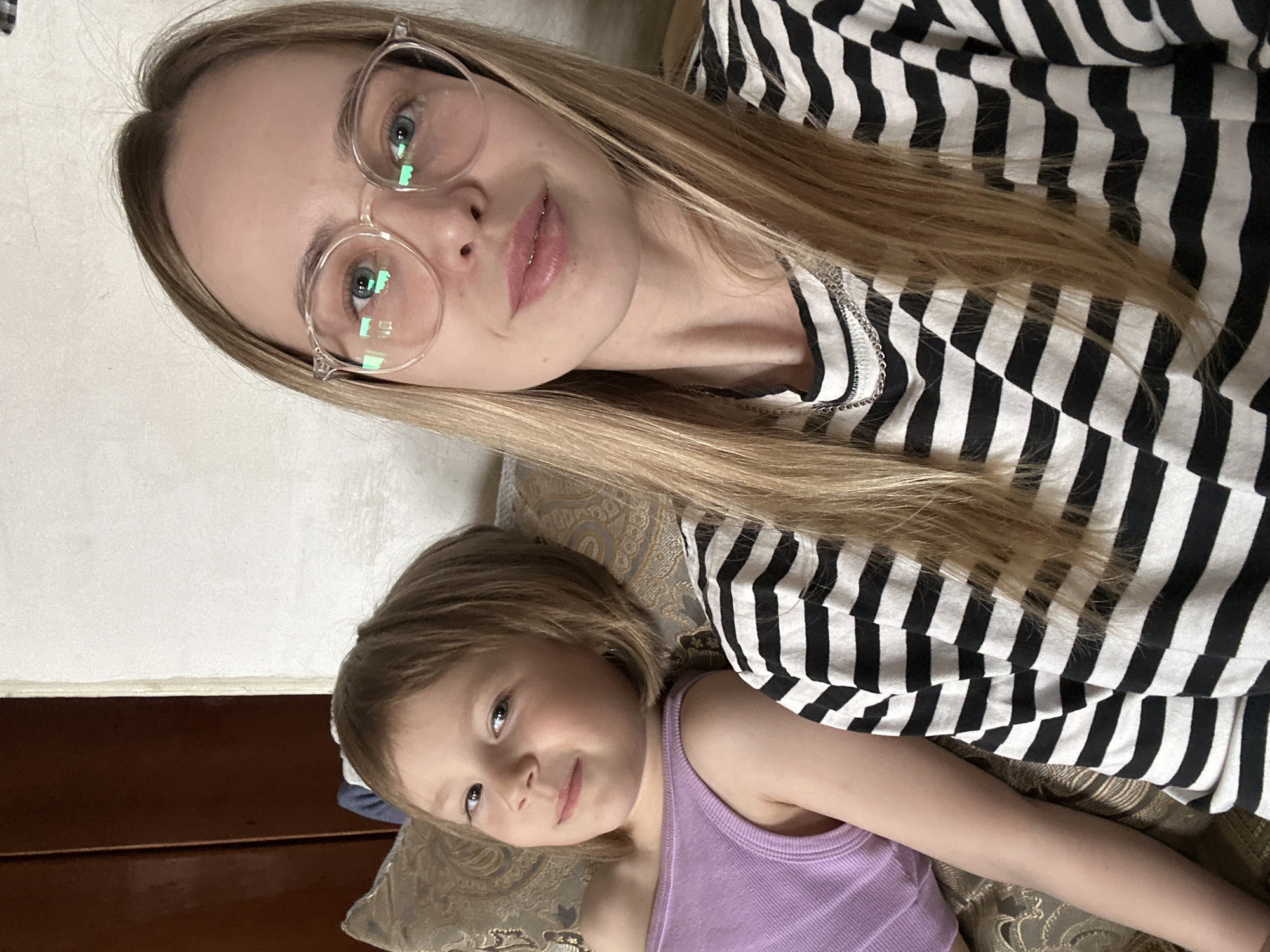 A woman and child are lying down in an interior space. The woman, who appears to be in her 20s, has long blonde hair and is wearing glasses and a black and white striped shirt. The child, a girl, has short brown hair and is wearing a lilac-colored tank top. Both are looking toward the camera, and the woman is holding the camera. They are lying on a patterned pillow or cushion. Part of a white wall and a brown wooden surface are visible in the background. The location is specified as Niva, Russia, though there are no location specific details or distinguishing marks to place the image in the city of Niva, Russia.