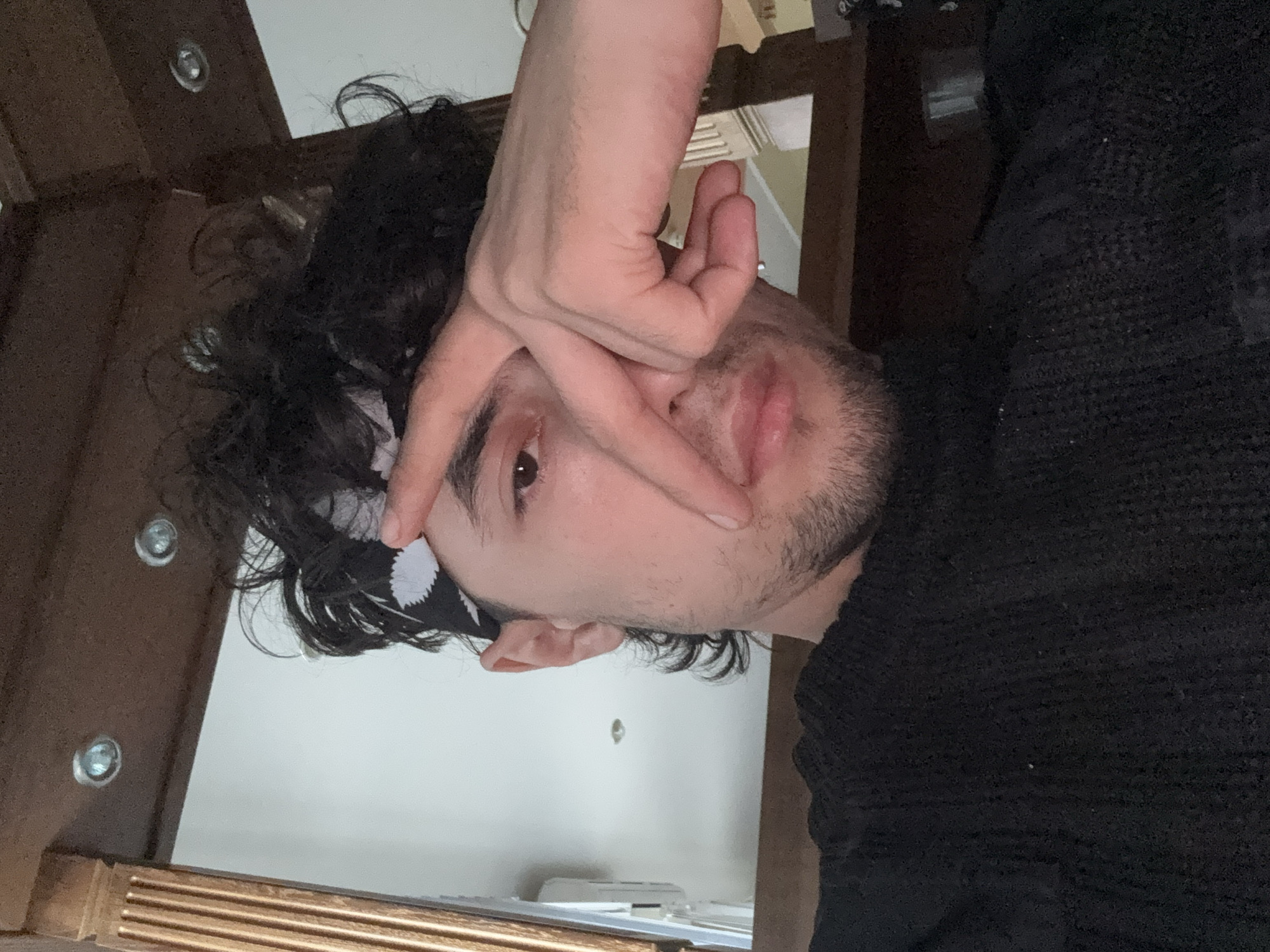 The image is a close-up, vertically oriented selfie of a person. The person has dark, curly hair, a light complexion, and is wearing a black sweater. A black and white patterned bandana is visible on their forehead. Their right hand is positioned over their face, with fingers obscuring part of their forehead and eye. The person's left eye is visible, looking towards the camera with a neutral expression. There is a hint of stubble on their face. The background appears to be indoors, with wooden furniture visible, suggesting a home or indoor setting. The lighting is soft, indicating it's likely daytime or an indoor setting with artificial light. There is no visible text in the image.
