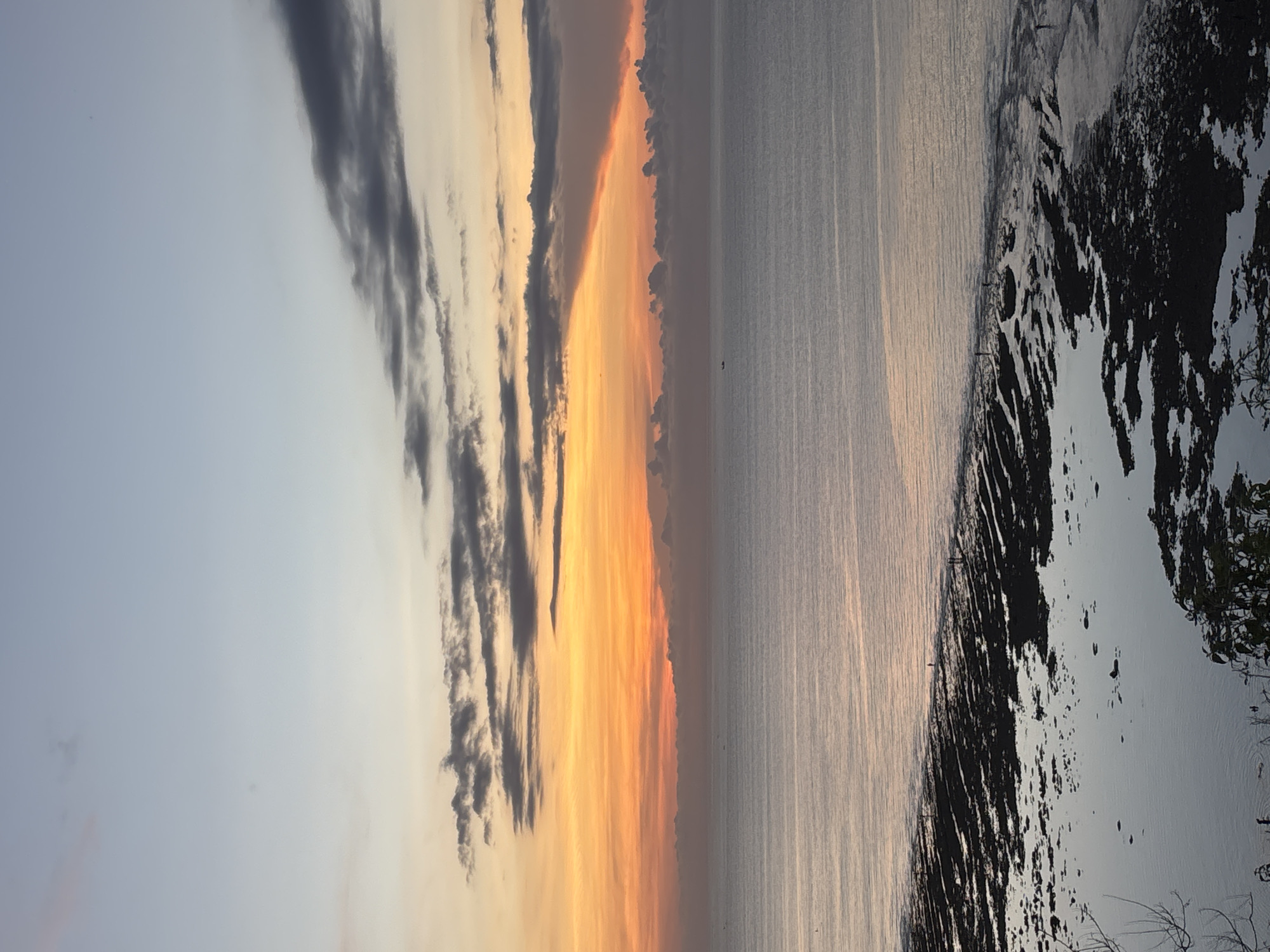 The image depicts a coastal sunset scene, likely in Kuta Selatan, Indonesia, given the context. The sky exhibits a gradient of color, transitioning from pale blue to fiery orange, with dark, elongated clouds. The ocean reflects the colors of the sky, creating shimmering patterns of light and shadow on the water's surface. The foreground reveals a shoreline, with a mix of dark, rocky areas and patches of lighter sand or shallow water. There is a thin line of vegetation present, suggesting that there are various types of plant life surrounding the area. The photograph captures a serene, natural environment at sunset.
