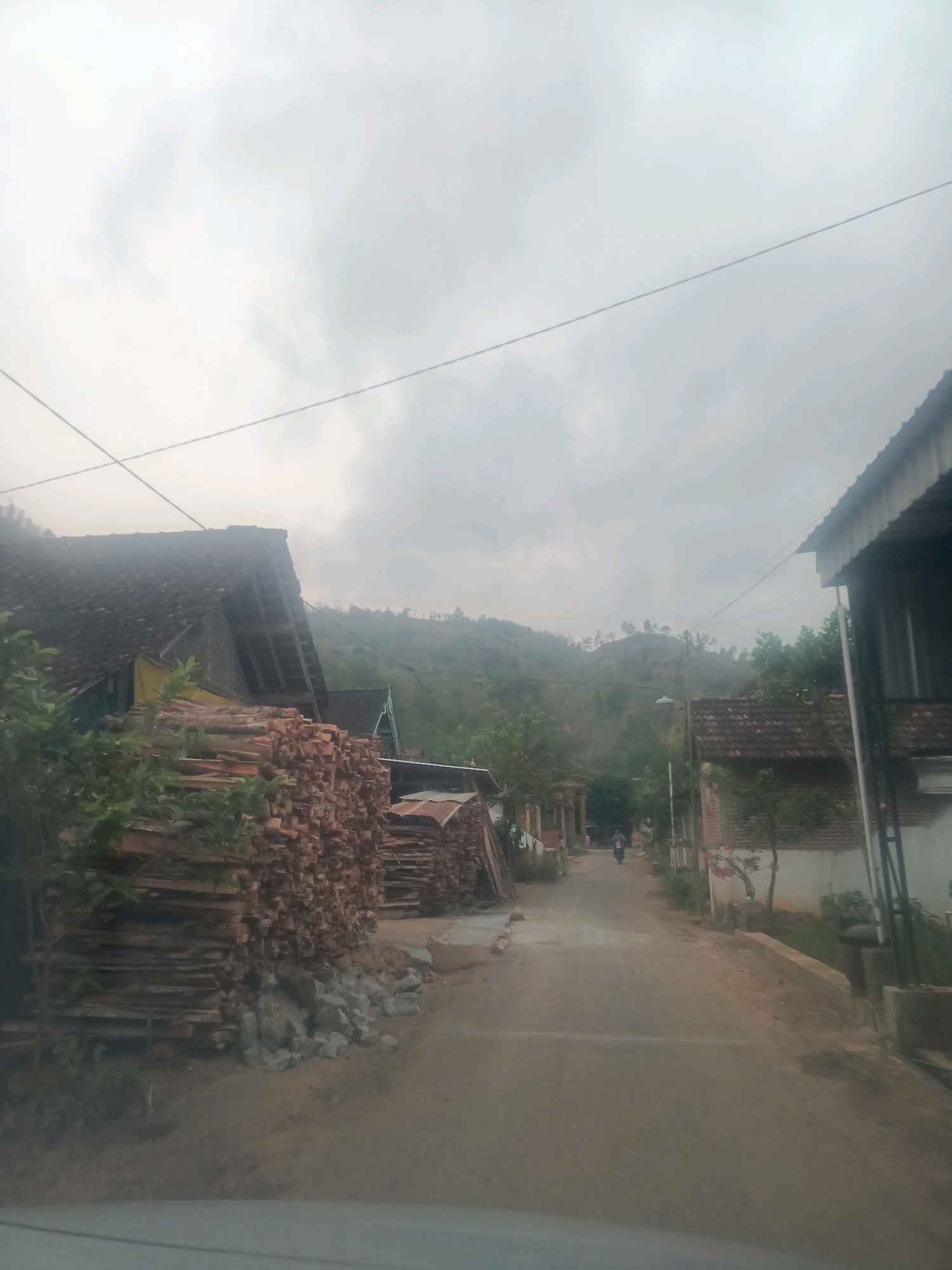 A rural road in Gandusari, Indonesia, is depicted under an overcast sky. On the left side of the road, a large stack of lumber is piled high, with a portion of a roofed structure visible behind it. To the right, another building with a corrugated metal roof and white siding is present. The road itself is unpaved and leads into the distance, where it curves around a hillside. A few trees and smaller buildings are scattered along the roadside. In the background, rolling hills covered in vegetation rise under the muted sky. A person on a motorcycle is visible on the road further ahead. The overall impression is one of a quiet, working village.