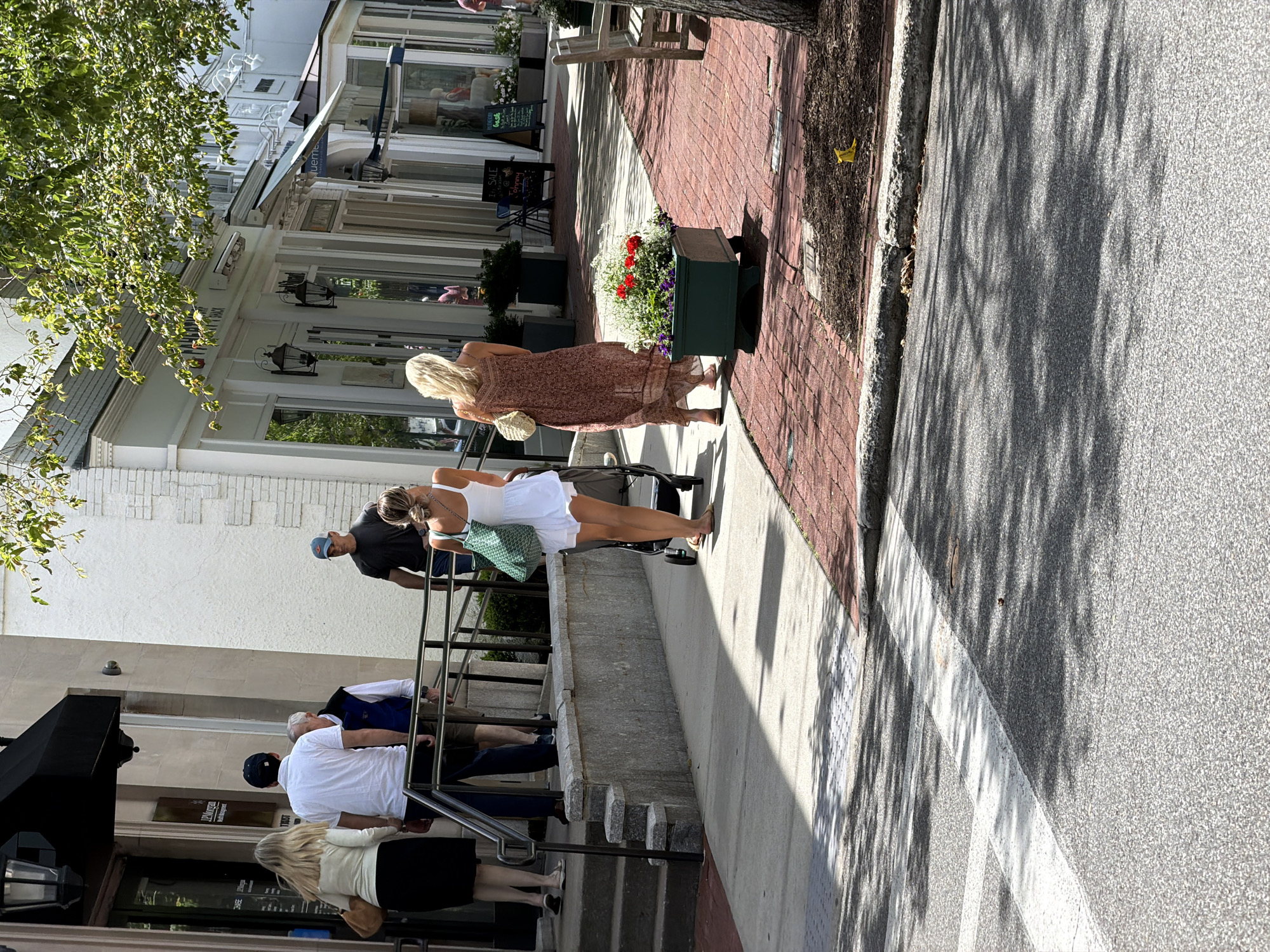 In the Village of Southampton, United States of America, on a sunny day, people are walking on a sidewalk next to a street. In the foreground, a crosswalk with white lines is visible on the asphalt road, with shadows of trees cast across it. To the right of the road is a red brick sidewalk, bordered by a concrete curb and then a strip of mulch with a yellow object, possibly a banana peel, near a tree.
On the sidewalk, two women are walking away from the camera. The woman on the left is wearing a long, patterned, reddish-brown dress and carrying a straw bag. The woman on the right is wearing a short white dress and carrying a green tote bag. She is pushing a stroller. Behind them, a group of people are ascending stairs leading to a building entrance. One man is wearing a dark t-shirt and a baseball cap, walking down the stairs. Another man in a white shirt and dark pants is walking up the stairs, followed by a man in a blue vest over a white shirt. A woman in a cream-colored top and a black skirt is also on the stairs. The buildings lining the sidewalk appear to be shops, with large windows and awnings. One building has a "JPMorgan Chase" sign. The environment is well-lit, suggesting daytime.