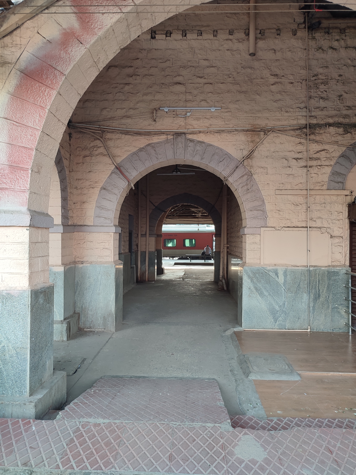 The image shows a view through a series of arched stone passages in a building, likely a train station platform in Unknown, Unknown. The arches are constructed from light-colored stone, with some having a darker gray base. The floors are a mix of concrete and patterned tiles. Through the arches, a red train car is visible. A person is sitting on a bench next to the train. The overall lighting suggests an indoor location with some natural light filtering through.
