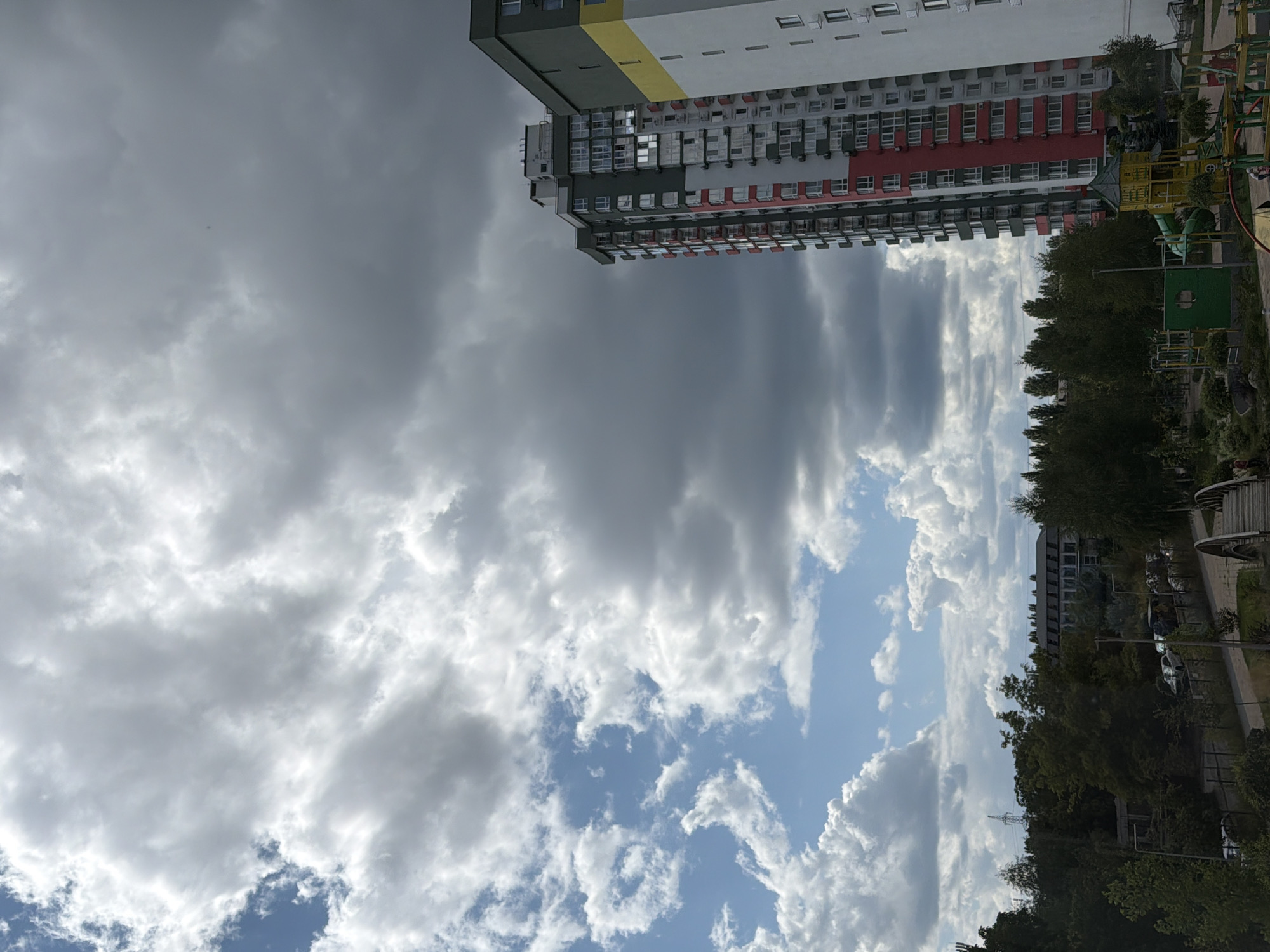 The image captures an urban landscape in Kyiv, Ukraine, dominated by a dramatic sky with both bright white cumulus clouds and darker, more ominous storm clouds. In the foreground, a modern, tall residential building with red and white accents stands prominently on the right side of the frame. Adjacent to it, another building with white and yellow sections is partially visible.
Below the buildings, a dense line of green trees forms a natural barrier. Beyond the trees, a low-rise building with a dark roof is visible, and further back, a few cars are parked along a road. A playground area with colorful equipment, including slides and climbing structures, is situated in the lower right portion of the scene, with a few small figures possibly present. A pedestrian bridge with an arched railing is visible in the lower right corner. The overall impression is one of an urban environment under a dynamic sky, with a mix of residential development and green spaces.