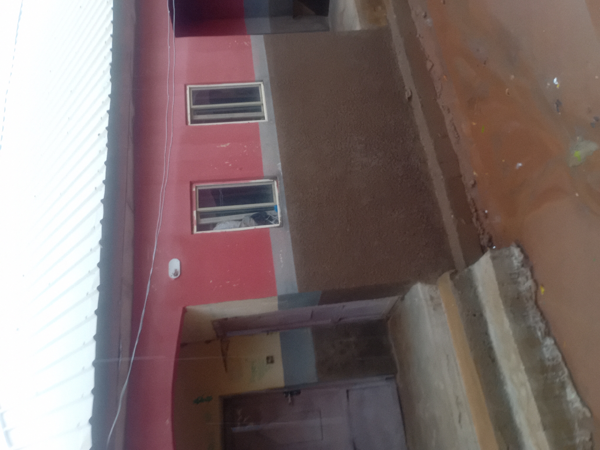 The image shows the exterior of a building with a red upper section and a brown lower section. There are two windows on the red section, each with white frames. A gray stripe separates the red and brown sections. To the left of the windows, there is an entrance with a purple door. The building has a white, corrugated metal roof. In front of the building, there are concrete steps leading up to the entrance. The ground in front of the building is a muddy, reddish-brown color, suggesting recent rain or damp conditions. The scene appears to be in a rural or semi-urban area, as indicated by the simple building construction and the natural ground cover. The location is specified as Jauro Yinu, Nigeria.
