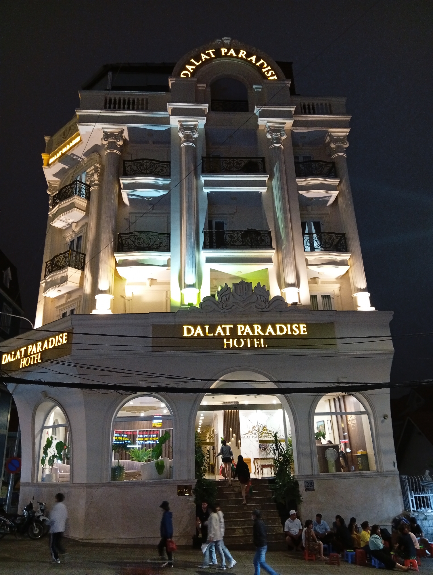 A nighttime shot of the Dalat Paradise Hotel in Dalat, Vietnam. The hotel is a multi-story building with a white facade and ornate architectural details, including columns and balconies. The name "DALAT PARADISE HOTEL" is illuminated in large, gold, neon letters on the front of the building, with a smaller sign above it that also reads "DALAT PARADISE HOTEL". The hotel entrance is at street level, with arched windows and a set of stairs leading up to the main doors. Inside the entrance, a reception area is visible, with plants and seating. Several people are walking around the hotel entrance and on the sidewalk in front of it. Some are walking up the stairs, while others are sitting on small stools on the sidewalk. The sky is dark, indicating it is nighttime, and there are no clear indications of the weather. The scene conveys a sense of arrival or departure at a hotel, with people coming and going.
