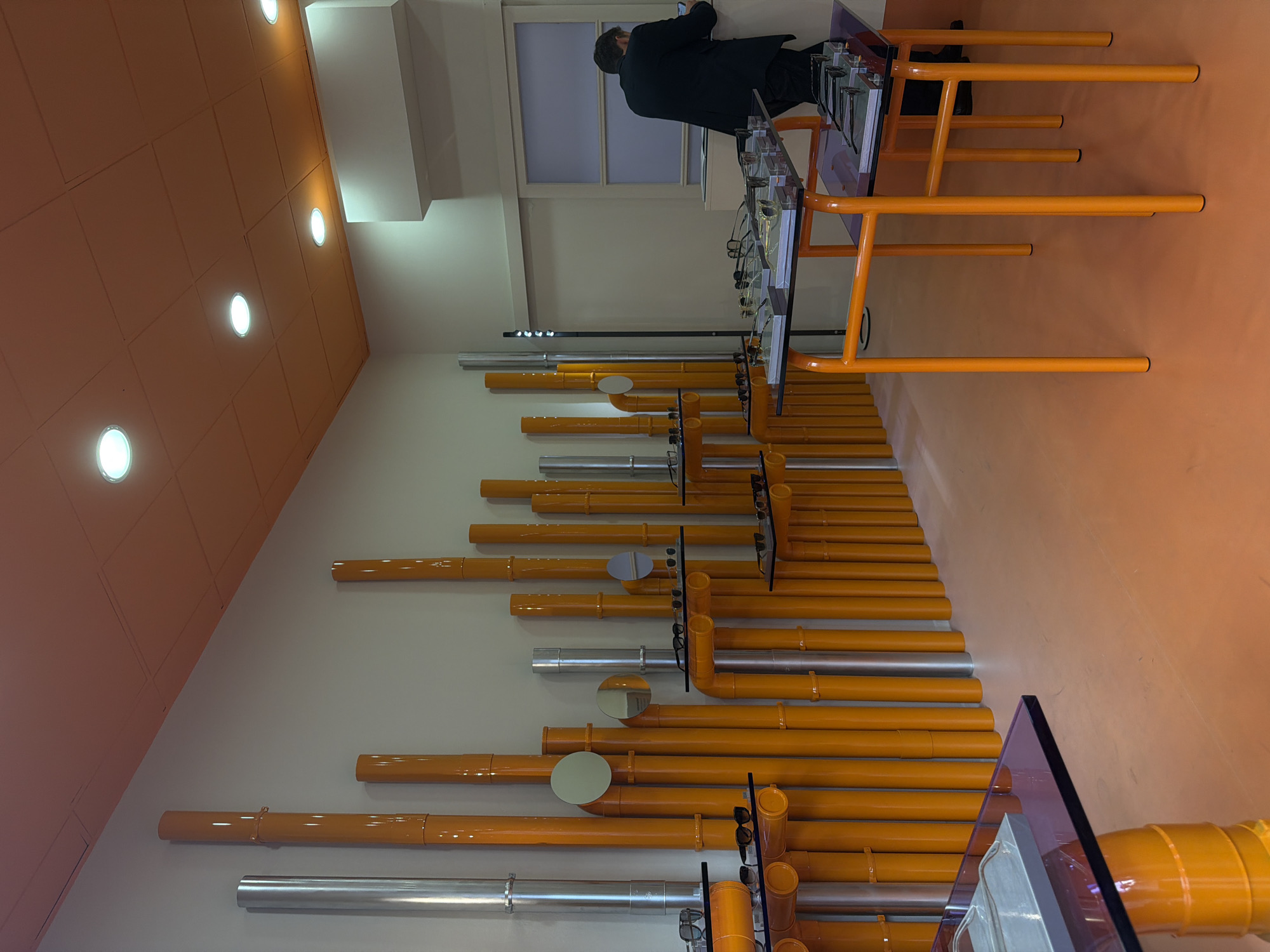 This image captures the interior of a modern retail or exhibition space, characterized by a distinctive design and vibrant color scheme. The scene is brightly lit by recessed circular spotlights embedded in an orange/ochre ceiling. The same orange/ochre color extends down one wall and across the floor, creating a continuous, immersive backdrop. The opposing wall is a clean off-white.
A prominent feature is a unique display system constructed from horizontal and vertical pipes or tubes mounted on the off-white wall. These pipes alternate between a glossy orange finish and a metallic silver finish. Interspersed among the pipes are circular, reflective elements, possibly mirrors or decorative discs, in silver or light gray. Numerous pairs of sunglasses are meticulously displayed on clear acrylic stands, attached to or placed on these pipe structures.
In the mid-ground, a person, viewed from behind, wears a dark suit or jacket and trousers. They are standing at an orange table with a clear glass top, which also displays several pairs of sunglasses. The person appears to be engaged with the display, possibly examining an item or using a mobile device. Behind them, a window with muted light or a frosted pane is visible. Another similar orange table with a glass top is partially visible in the foreground, hinting at more display space. The tables feature sturdy, angular orange legs.
The overall atmosphere is contemporary and well-organized, with a striking contrast between the bold orange elements and the minimalist white and silver tones. No discernible text is visible in the image. Given the provided context, the location is Milan, Italy, suggesting a design-forward commercial or exhibition setting.