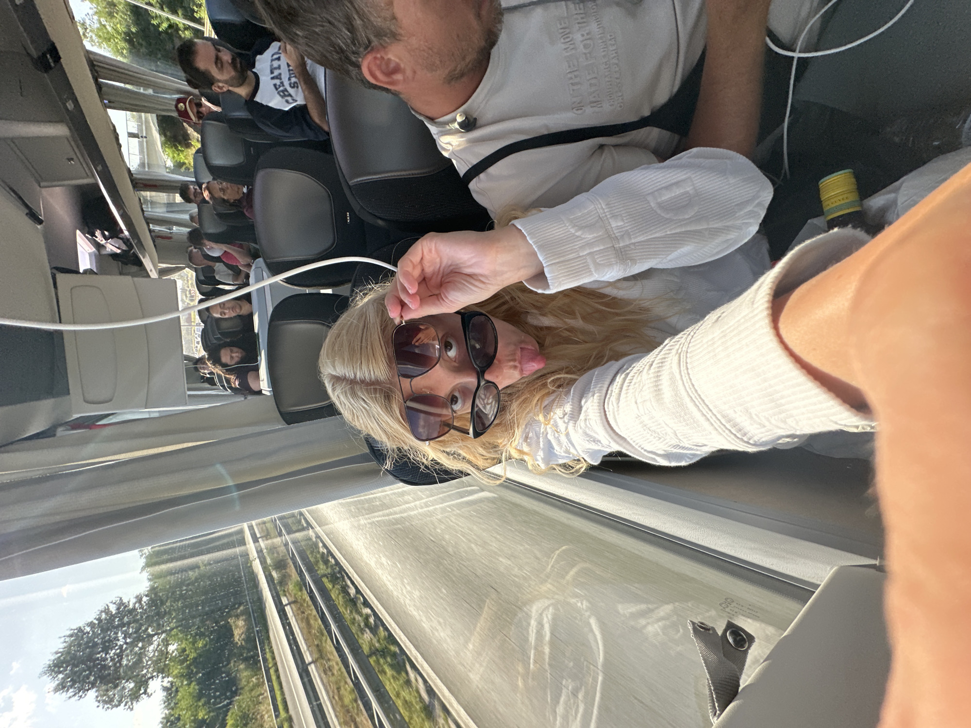 A blonde woman is taking a selfie on a bus. She is wearing a white top and has two pairs of sunglasses on her face, one pushed up onto her forehead. She is sticking her tongue out at the camera. In the background, other passengers are seated on the bus, and the window shows a view of trees and a highway. The bus is traveling on a highway, likely near Deinze, Belgium, given the context. The lighting suggests it is daytime.