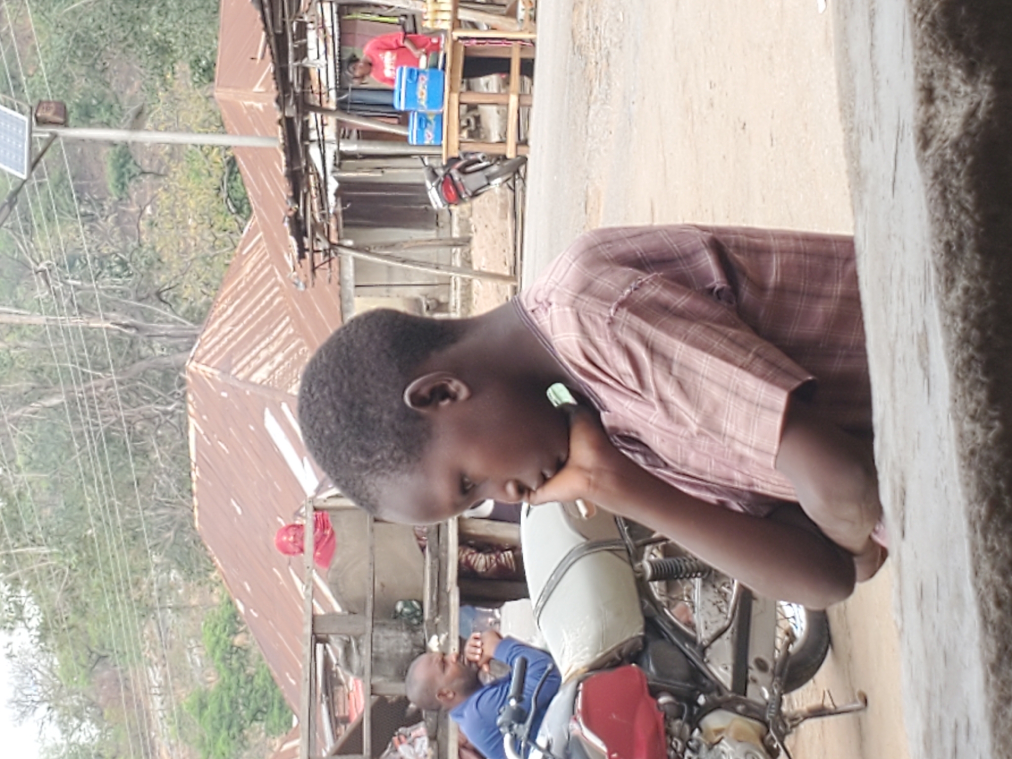 A young boy in a plaid shirt sits with his face resting on his hand while straddling a motorcycle. A man in a blue shirt is also on a motorcycle behind him. In the background, shops with corrugated metal roofs line the street, some displaying merchandise. A solar panel stands tall amidst utility lines. The location appears to be a street in Mararaba, Nigeria.