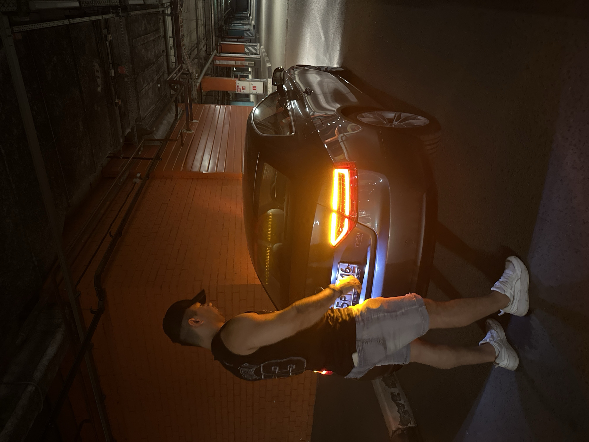 In a dimly lit, multi-story parking garage, a man stands next to a dark-colored coupe. The man, wearing a black sleeveless shirt, denim shorts, a black baseball cap, and white sneakers, is facing away from the camera and looking towards the rear of the vehicle. The car's taillights are illuminated with a bright orange glow, reflecting off the surrounding concrete and brick walls. The license plate of the car is partially visible, displaying Cyrillic characters and numbers that appear to be "5PM 16". The garage features exposed pipes and conduits on the ceiling, characteristic of an urban infrastructure. The overall atmosphere is one of a typical evening scene in a city parking facility, possibly in Kazan, Russia, given the license plate details.