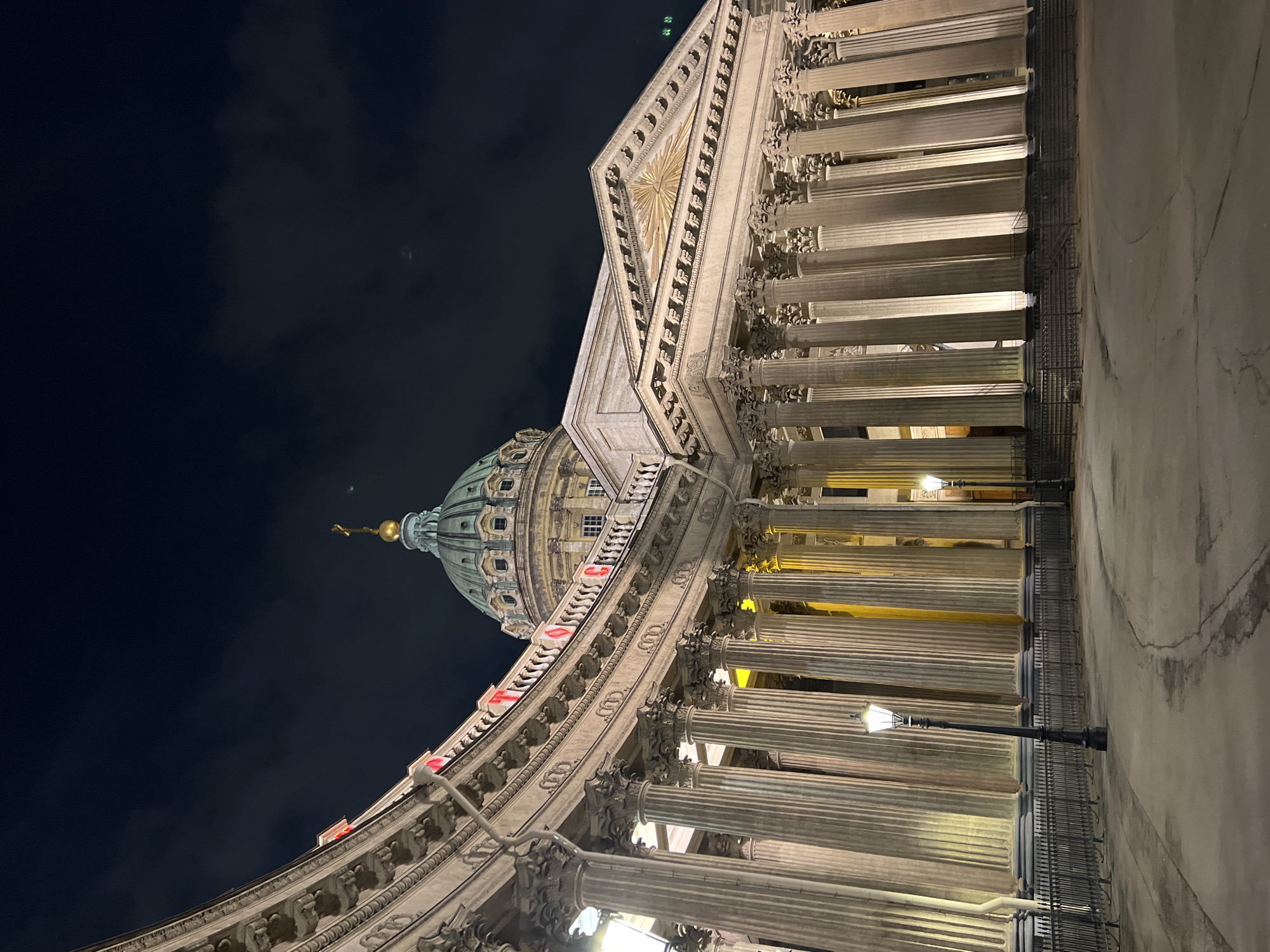 The image captures a nighttime view of the Kazan Cathedral in Saint Petersburg, Russia. The building's architecture is predominantly visible, featuring a long colonnade constructed of numerous Corinthian columns, which are illuminated from below. The facade leads to a large dome, topped with a golden spire. The sky is a dark shade of blue or black. The street beside the columns is visible and contains a historic-style street light. The perspective is unusual, looking up at an angle towards the dome.