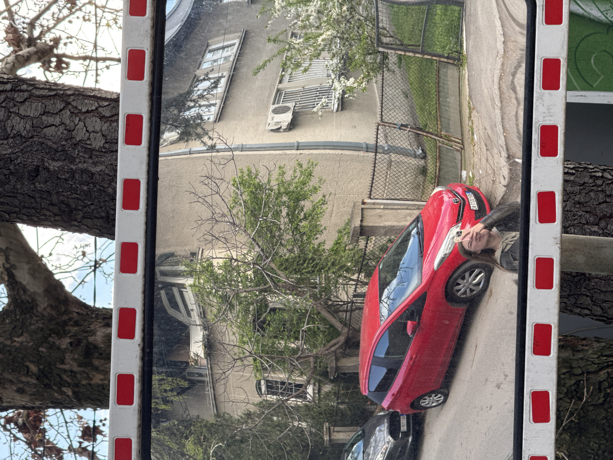 The image shows a reflection in a street mirror, likely used to improve visibility at a blind intersection. The reflection features a residential building with several windows and an air conditioning unit, a red car, a tree with some green foliage, and a person (likely a young woman) taking a photograph with a mobile phone. The mirror itself has a white frame with red rectangular reflectors. A tree branch is visible to the left of the mirror frame. It is plausible the photo was taken on a street in Vozdovac Urban Municipality, Serbia, based on the license plate visible on one of the cars.