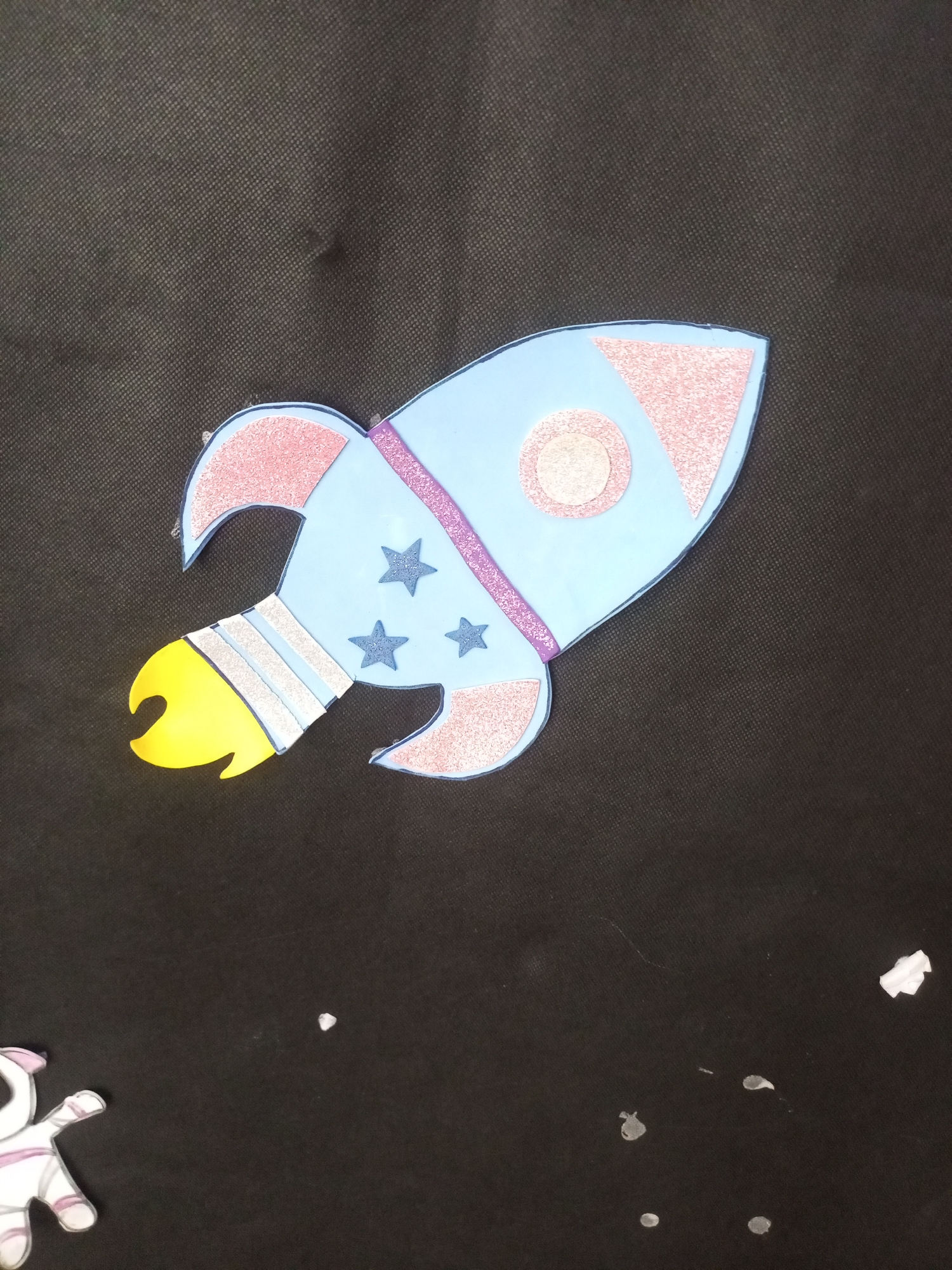 The image is a close-up, overhead shot of a handmade rocket craft lying on a dark, textured surface. The rocket is light blue with pink glitter accents on the fins and a circular window. It has three dark blue stars on its body and a yellow flame coming out of the engine. Silver and blue bands adorn the engine section. A small portion of a white and pink astronaut figure is visible in the lower left corner. The background is a dark, fabric-like material with a faint speckled pattern. There are a few small, white, irregular shapes scattered on the background, possibly paper scraps or debris. The location of Santa Ana, Venezuela is not depicted or suggested by any elements within the image.
