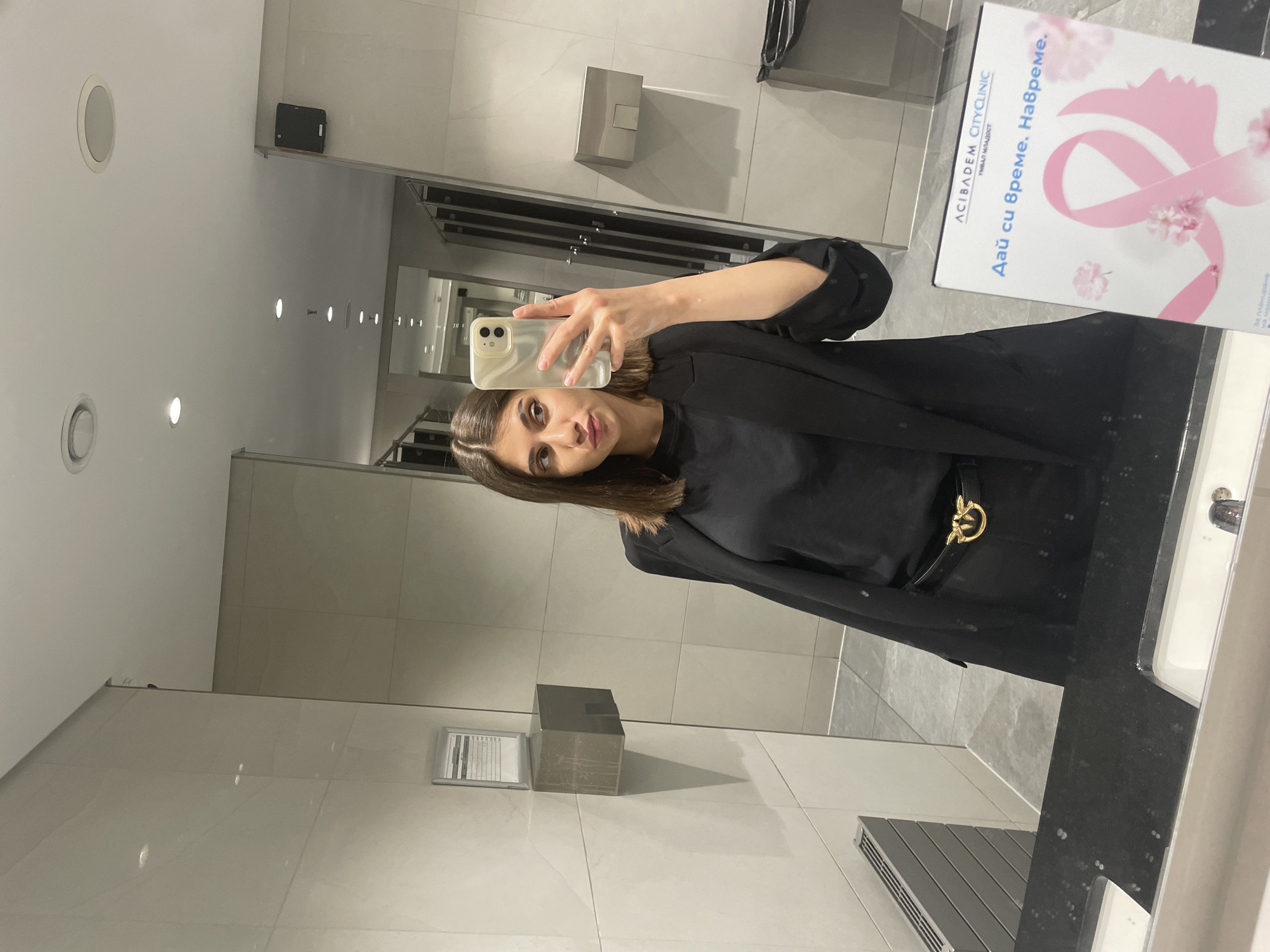 A woman is taking a selfie in a bathroom. She is holding a smartphone in her right hand. She is wearing a black shirt and a black blazer. There is a black belt with a gold buckle on her waist. The bathroom has white tile walls. There is a mirror in front of her. There is a sink and a counter to her right. There is a hand sanitizer dispenser on the wall. There is a poster on the counter with the words "ACIBADEM CITY CLINIC" and "Дай си време. Навреме." and a pink ribbon. It is possible the location is Sofia, Bulgaria, based on the Bulgarian text on the poster.