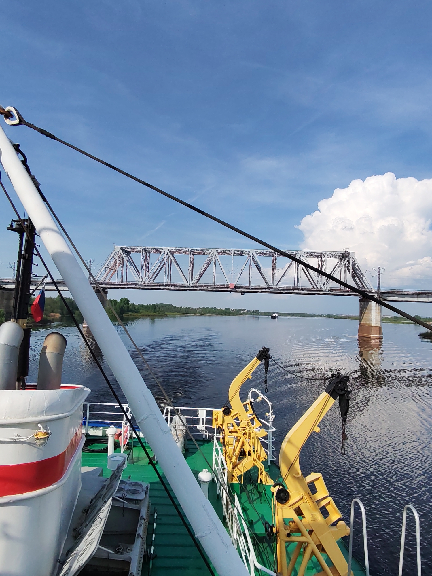 View from a boat, possibly a research or utility vessel, on a wide river in the city of Нифантово, Russia. In the foreground, there is equipment on the deck of the boat including yellow cranes, and a white painted structure with a red stripe. A Russian flag is visible attached to a pole. A large metal truss bridge spans the river in the middle distance with a secondary bridge adjacent to it. A small barge is moving along the water in the middle of the scene. The river reflects the sky and bridge. In the background is a blue sky with wispy clouds and a large cumulus cloud. Green vegetation is visible along the riverbank.