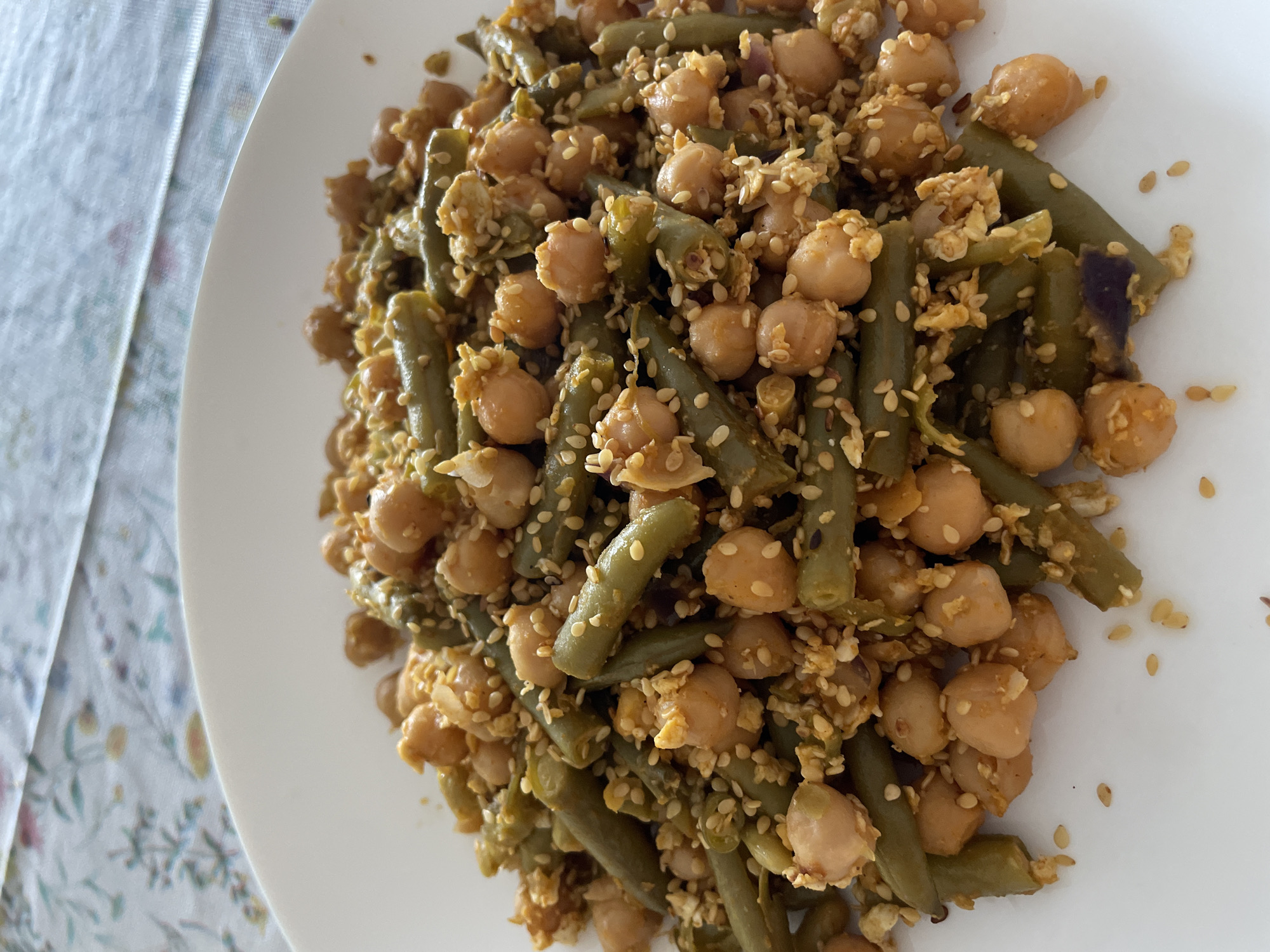 The image shows a close-up of a white plate filled with a dish consisting of green beans and chickpeas, seasoned with sesame seeds and possibly other spices. The plate appears to be placed on a tablecloth with a floral pattern. The location, Senden, Germany, is not discernible from the visual content of the image.