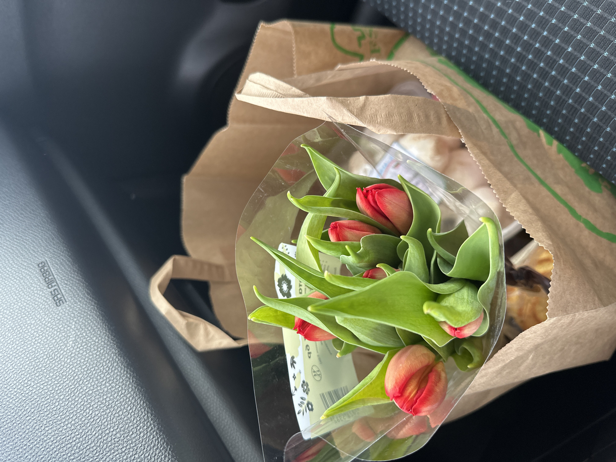 The scene is an interior shot within a vehicle, likely a car, where a brown paper bag sits on the passenger seat. The bag contains a bouquet of red tulips encased in clear plastic wrapping. Some baked goods are partially visible within the bag, alongside the flowers. The car's interior is dark, with a textured gray dashboard and a patterned seat in the background. The inscription "SRS AIRBAG" is visible on the dashboard. There is no evidence in the image to establish that the location is in the city of Burgas, Bulgaria.