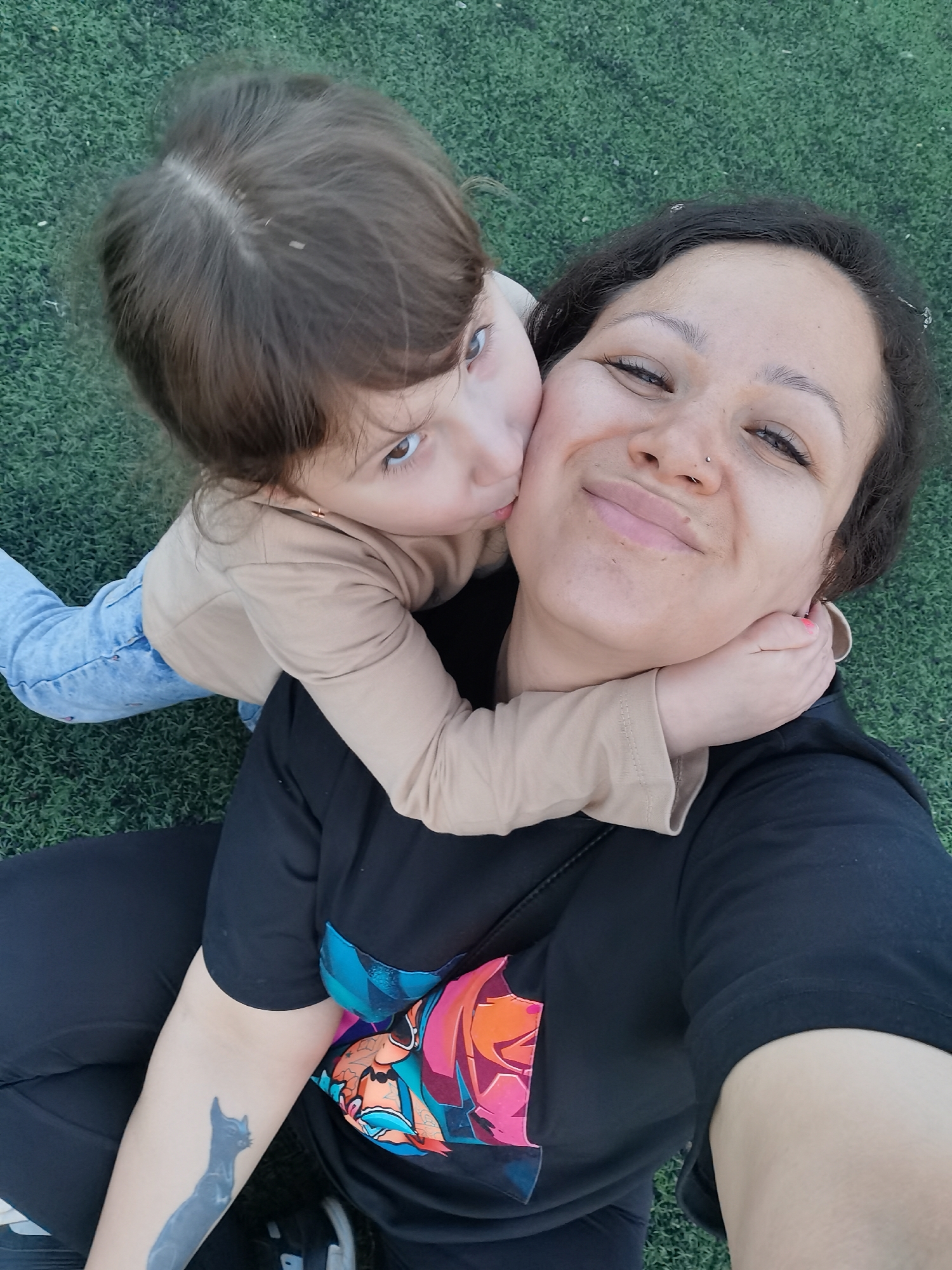 A young child with light brown hair and a beige long-sleeved shirt embraces a woman. The child's left cheek is touching the woman's right cheek as if giving a kiss. The woman is smiling, and has black hair and wears a black t-shirt with a colorful design on the left side of the chest. A black cat tattoo is visible on her left forearm. Both are positioned on green, artificial turf. The photo was taken outdoors and appears to have been taken in the city of Chaikovsky, Russia.
