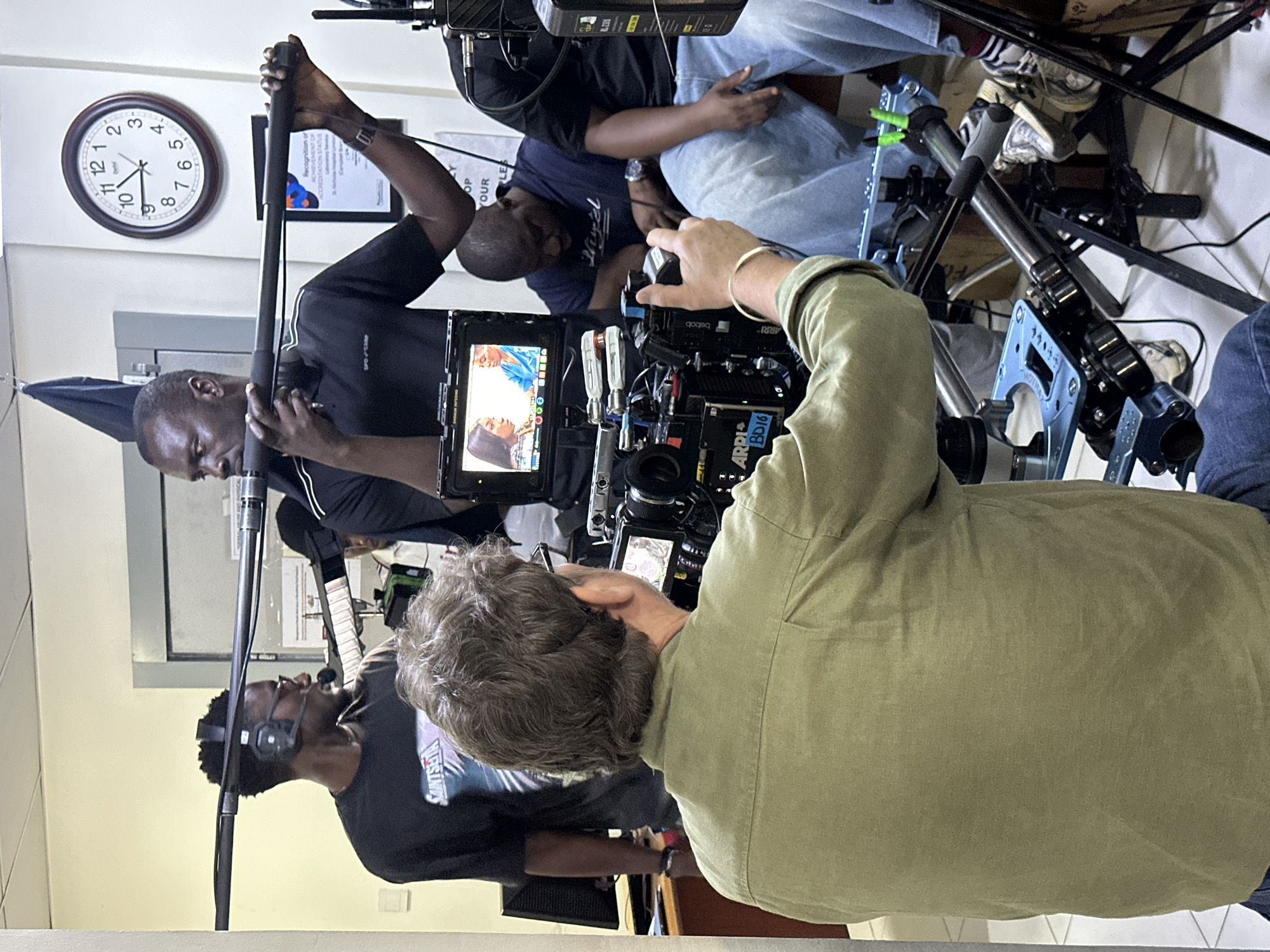 The image shows a film production scene. There are at least four people involved. One person is operating a professional camera, possibly an ARRI model (as indicated by "ARRI" and "B016" on the camera). Another person is holding a boom microphone. There is a monitor showing a recording of what appears to be an interview with two people. The location appears to be inside a building, as suggested by the wall and clock. The building looks like a clinic in Lagos, Nigeria. Further analysis of objects such as documents or branding could potentially corroborate the location.