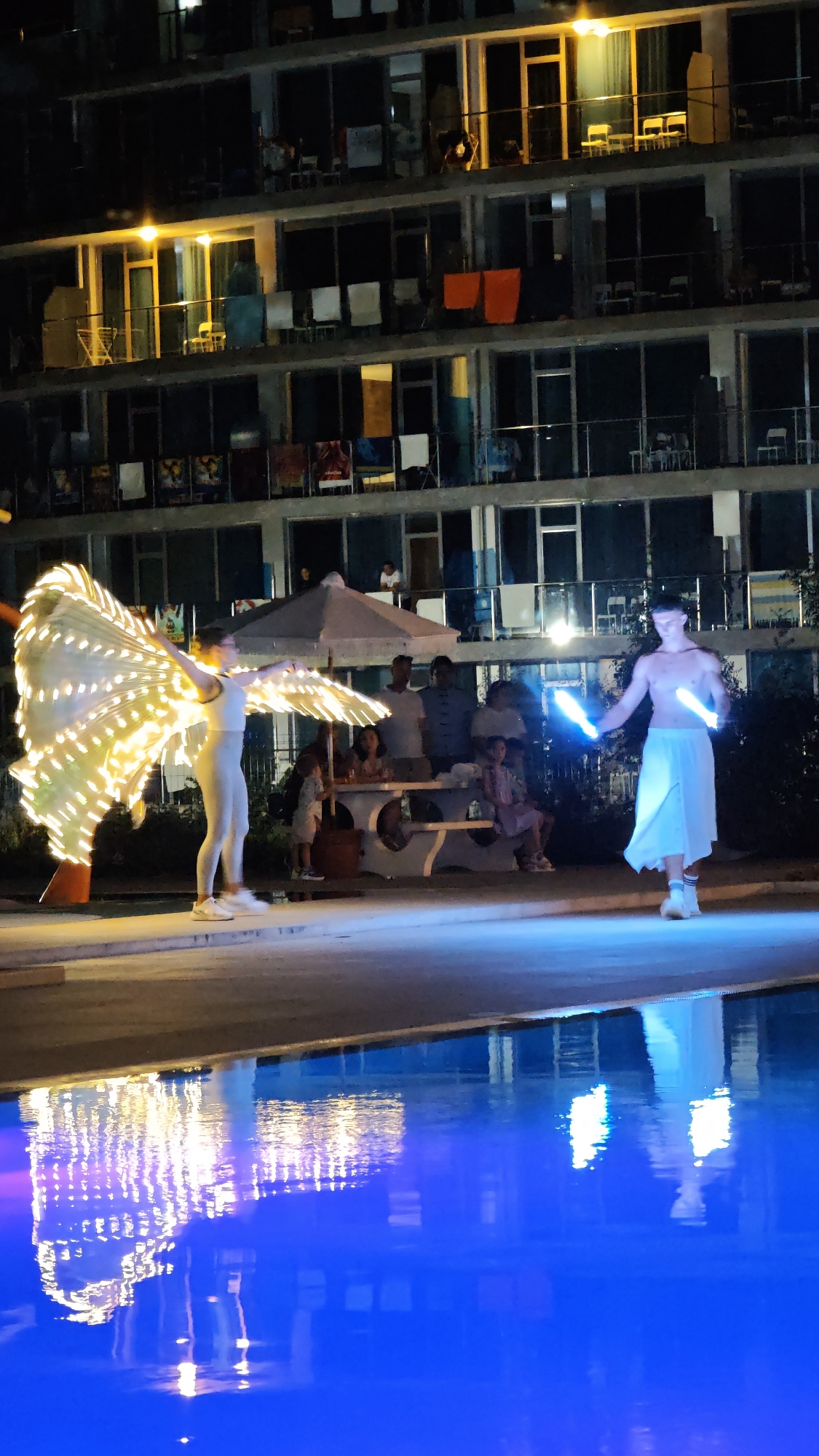 In Kiten, Bulgaria, a night scene unfolds with a performance by two individuals near a swimming pool. On the left, a person in a light-colored outfit wears a large, illuminated wing-like prop, shimmering with many small, bright lights. To the right, another person, shirtless and wearing a light-colored skirt, holds two glowing blue sticks. In the background, an audience of several people, including children, observes the performance from behind a picnic table, with an umbrella providing shade. Behind them, a multi-story building with balconies and illuminated windows stands as the backdrop, with laundry visible on some balconies, suggesting a residential or hotel setting. The swimming pool in the foreground reflects the lights and activity, creating a vibrant and dynamic atmosphere.