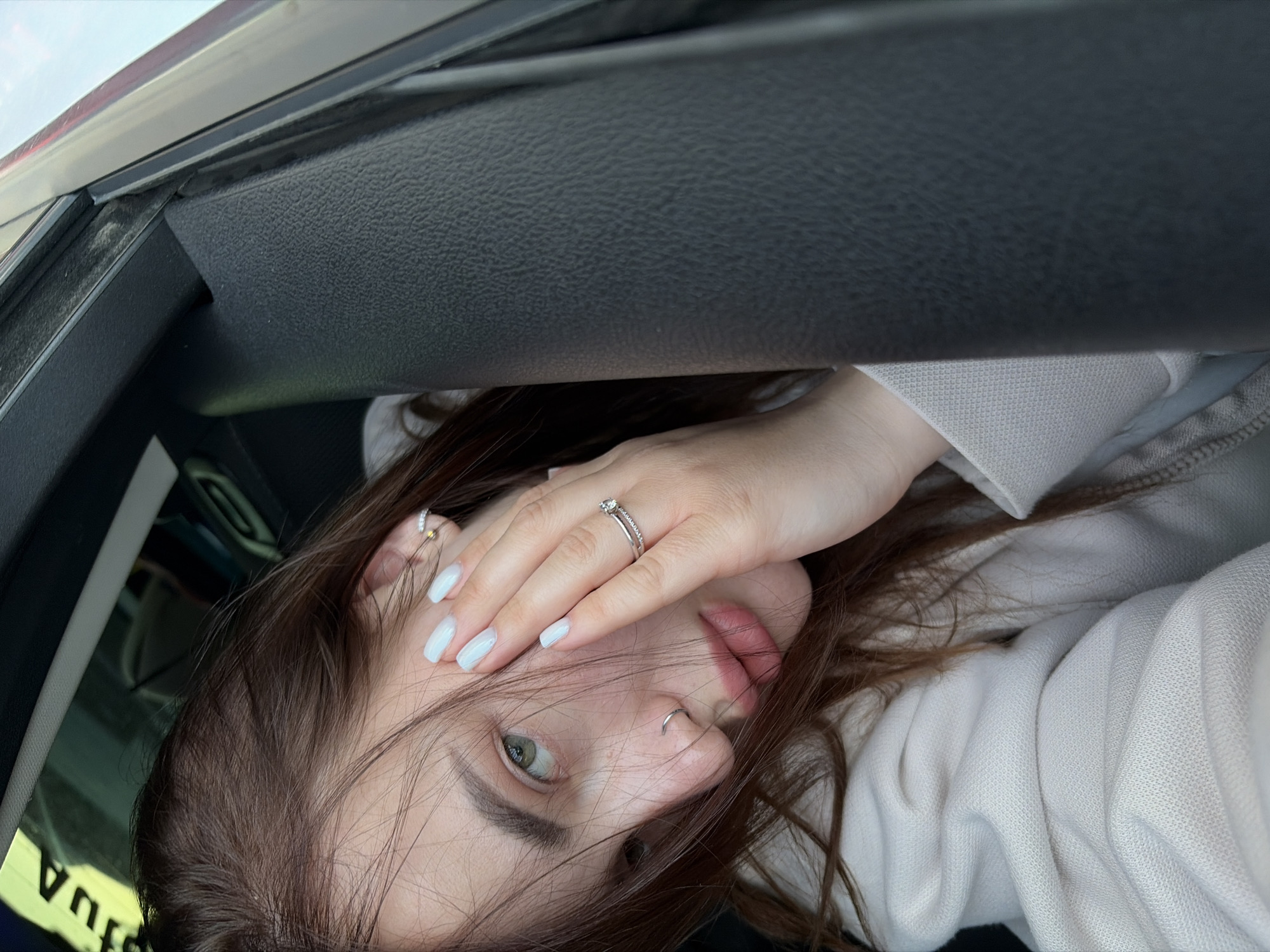 A young woman with light skin, brown hair, and green eyes is posing for a selfie inside a car. She is wearing a white hoodie and has a nose ring. Her nails are painted light blue, and she is wearing a ring on her left ring finger. She is holding her hand near her face, partially covering her eye. The car interior is visible, including the dashboard and window frame. The location is not identifiable as Krasnodar, Russia from the image alone.