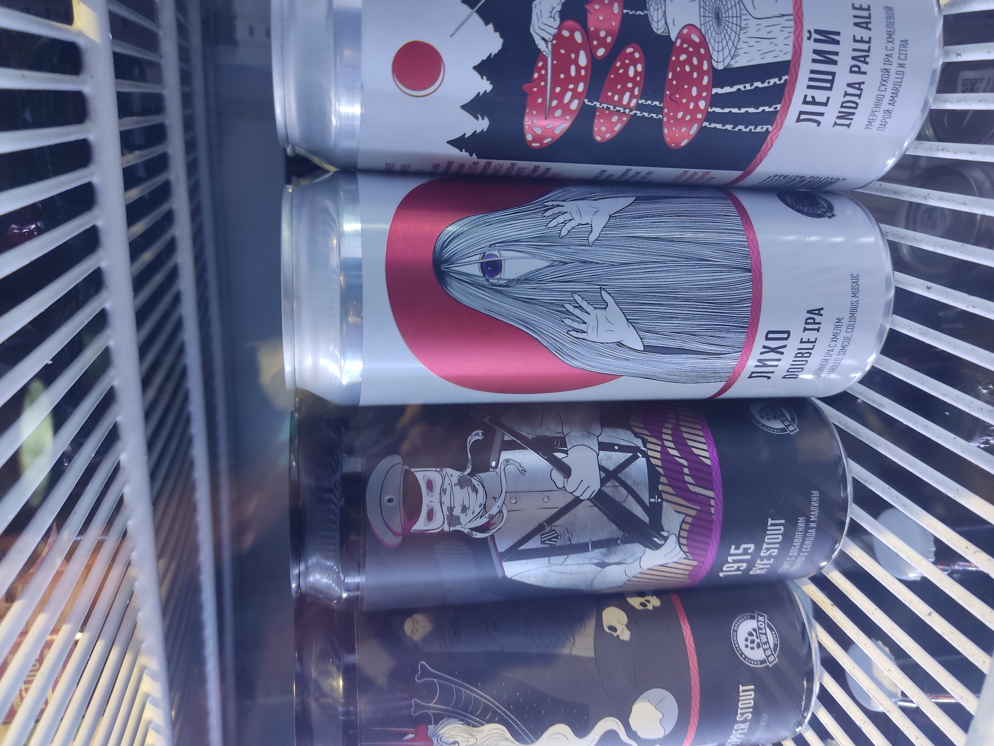 The image shows four cans of craft beer stacked vertically inside a refrigerated display unit, likely in a retail setting. The top can is labeled "ЛЕШИЙ INDIA PALE ALE," indicating it is an India Pale Ale. The second can is labeled "Лихо DOUBLE IPA," a Double IPA. The third can is labeled "1915 RYE STOUT," suggesting it is a Rye Stout. The fourth can is partially visible and appears to be a stout. All the can labels are in Russian, and the information provided with the image indicates this is likely from Pskov, Russia.
