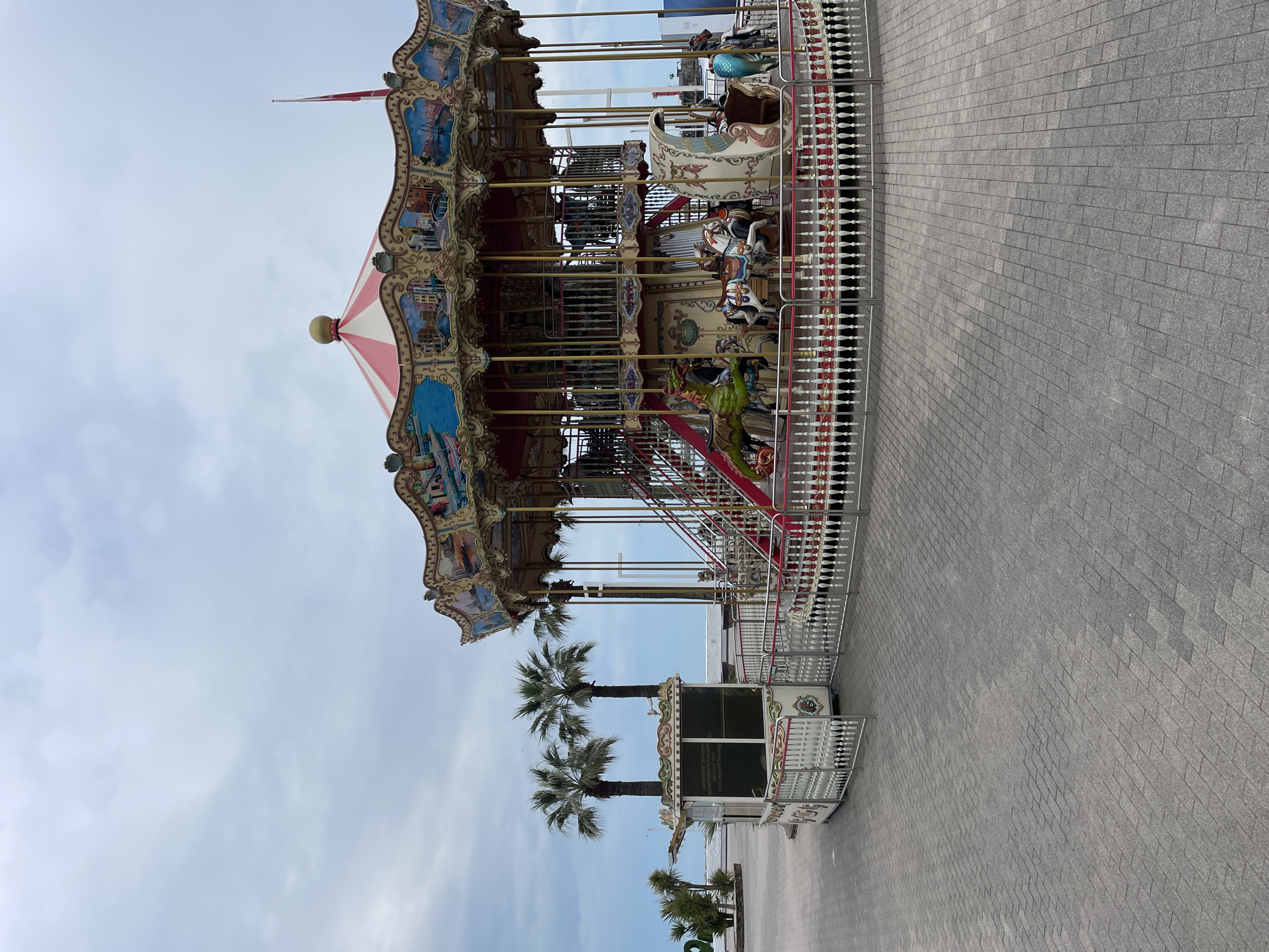 The image shows a two-tiered carousel with a striped red and white roof. The carousel is decorated with ornate paintings and features various animal figures. It is surrounded by a white picket fence and paved with grey bricks. Several palm trees are visible in the background, along with a small, decorated booth, possibly for ticket sales. The sky is overcast with light grey clouds. Given the presence of palm trees and architecture, the location is likely Sochi, Russia.