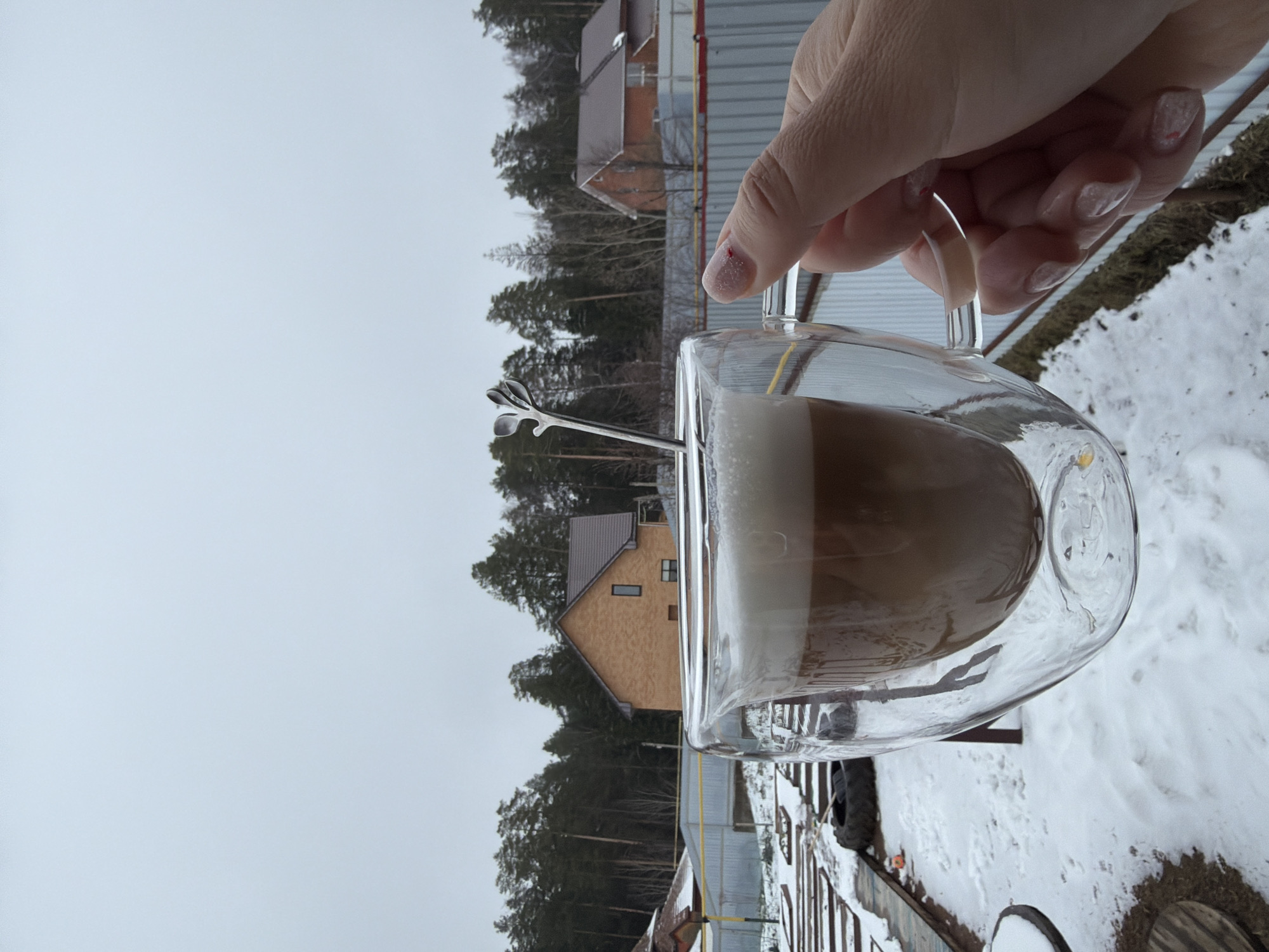 A human hand holds a glass mug filled with a coffee drink, likely a latte or cappuccino, with visible layers of coffee and milk foam. A decorative silver spoon is inserted into the drink. The background shows a residential area with houses surrounded by trees, suggesting a suburban or rural setting. The ground is covered in snow, indicating a winter season. The overall scene appears to be taken outdoors on a cold day, with a pale sky. It is impossible to verify that this is a city located in Chaikovsky, Russia.