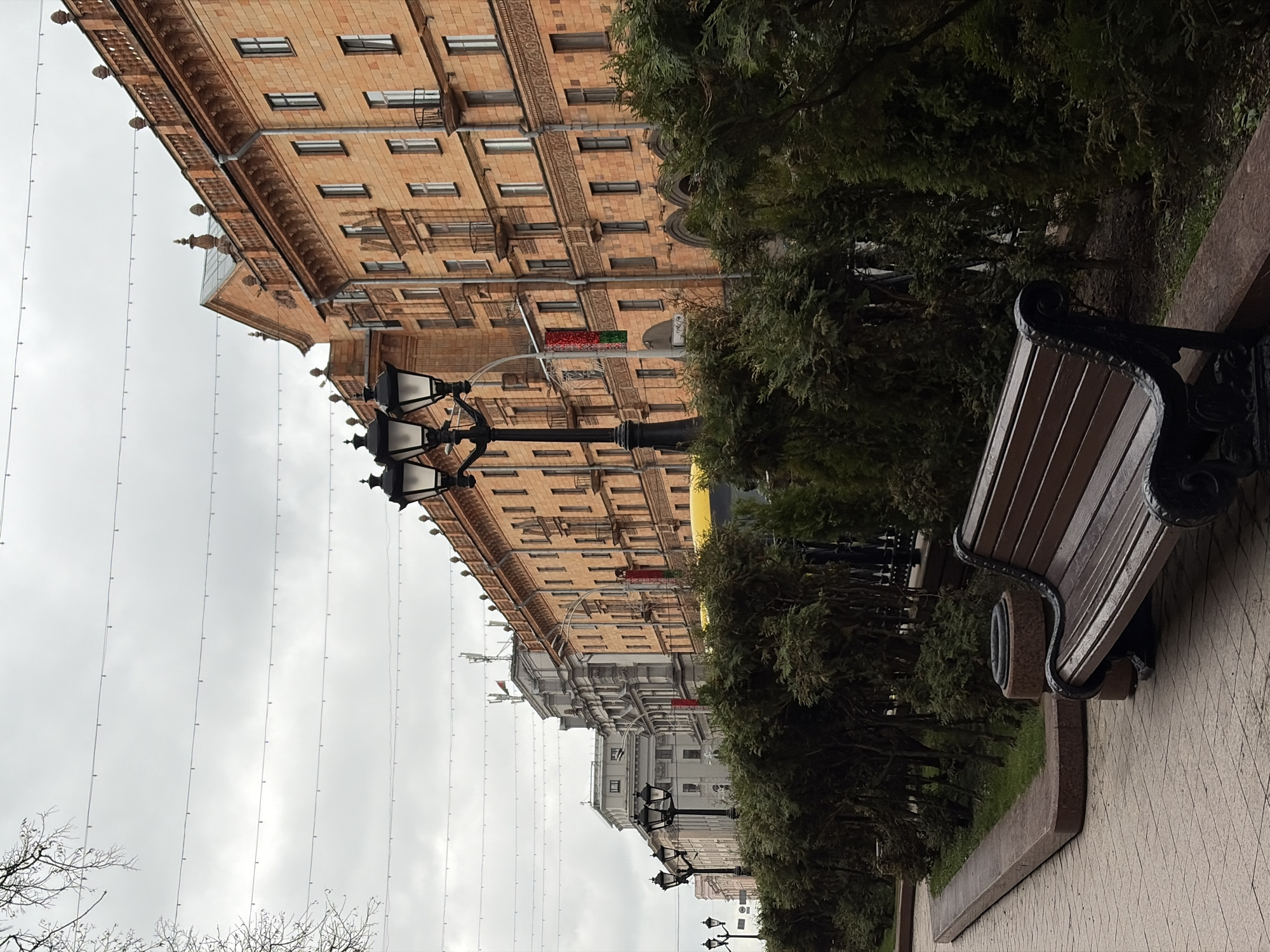 A street scene in Minsk, Belarus, featuring a large orange building lining the street with repetitive windows and ornate details, street lamps, evergreen trees planted in the median of the sidewalk, and a wooden bench placed on the edge of the walkway. The sky is cloudy and a line of wires spans overhead.