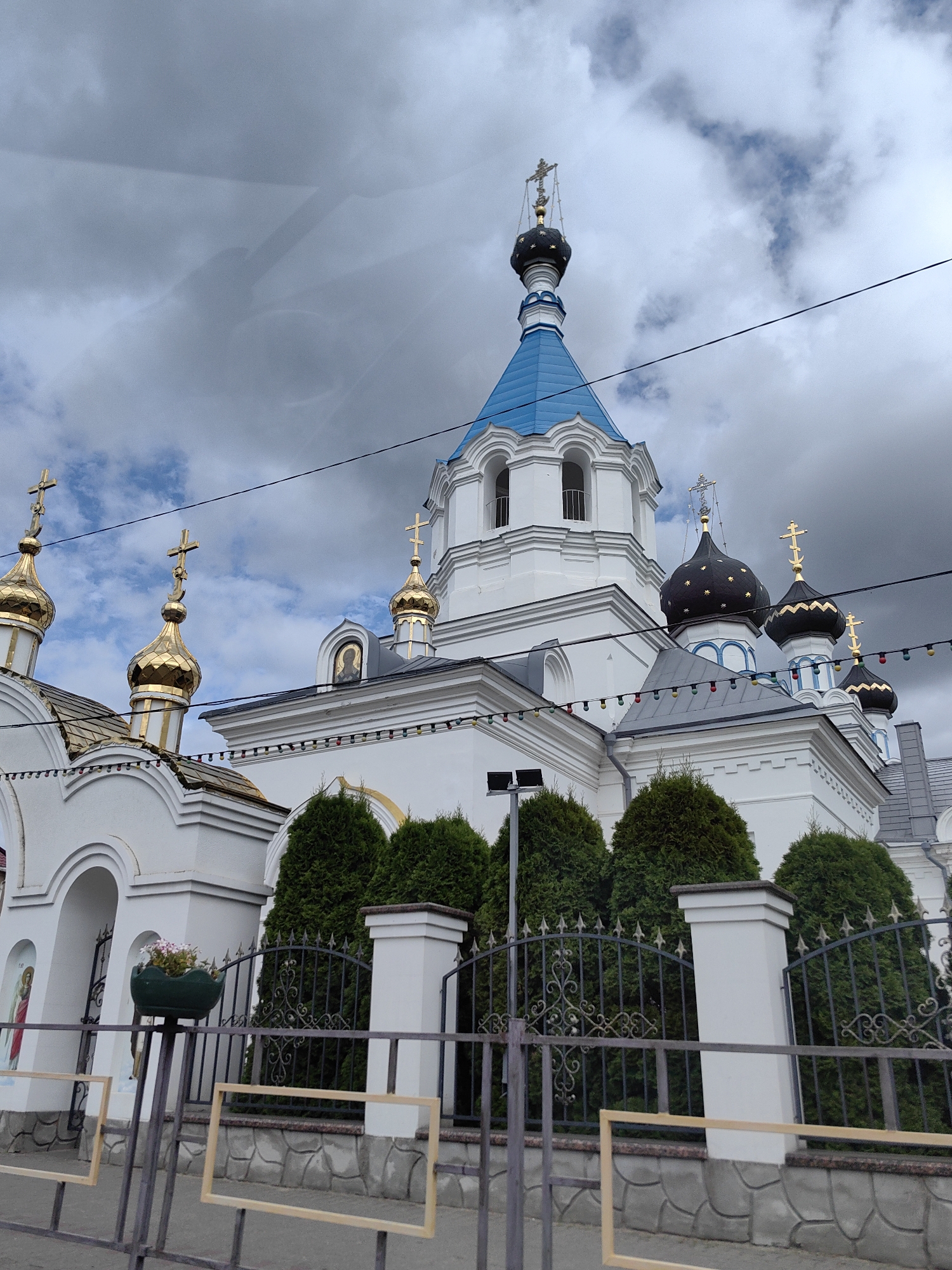 The image shows a white Orthodox church with multiple golden domes and one blue dome, located in Pastavy, Belarus. The church has intricate architectural details, including arched windows and decorative elements. In front of the church, there is a black metal fence and several green cypress trees. The sky is cloudy and overcast. There are also some colorful string lights decorating the building. Two empty wooden frames are visible in the foreground, attached to the fence.