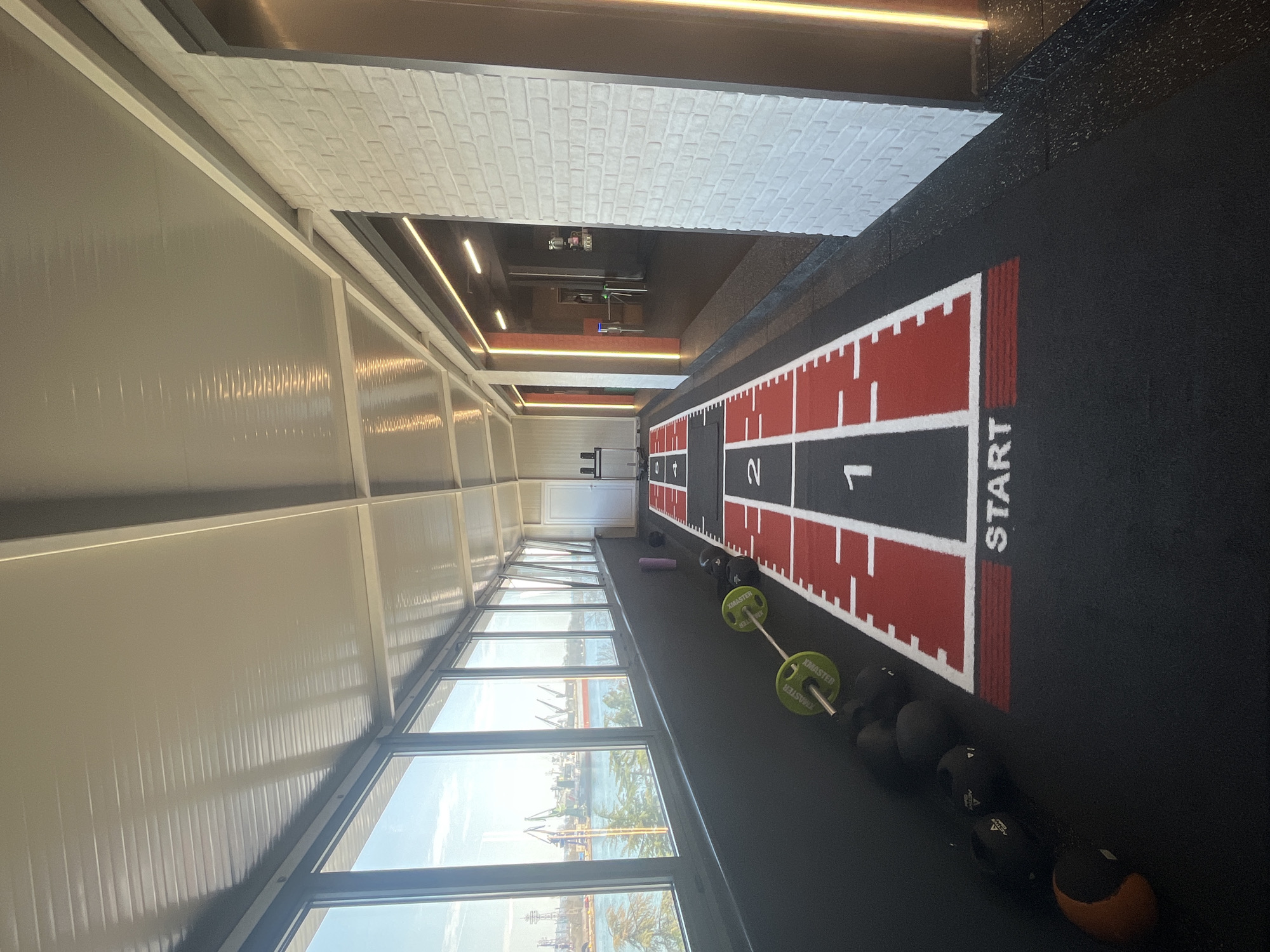 The image shows the interior of a gym located in the city of Unknown, Unknown. The gym has a running track marked with "START" and numbered sections. Exercise equipment such as medicine balls and a barbell with weights are placed along the track. The walls are a mix of white brick and glass, offering a view of a harbor with ships and industrial structures. The room is well-lit with both natural light from the windows and artificial lighting fixtures. There are no people visible in the image.