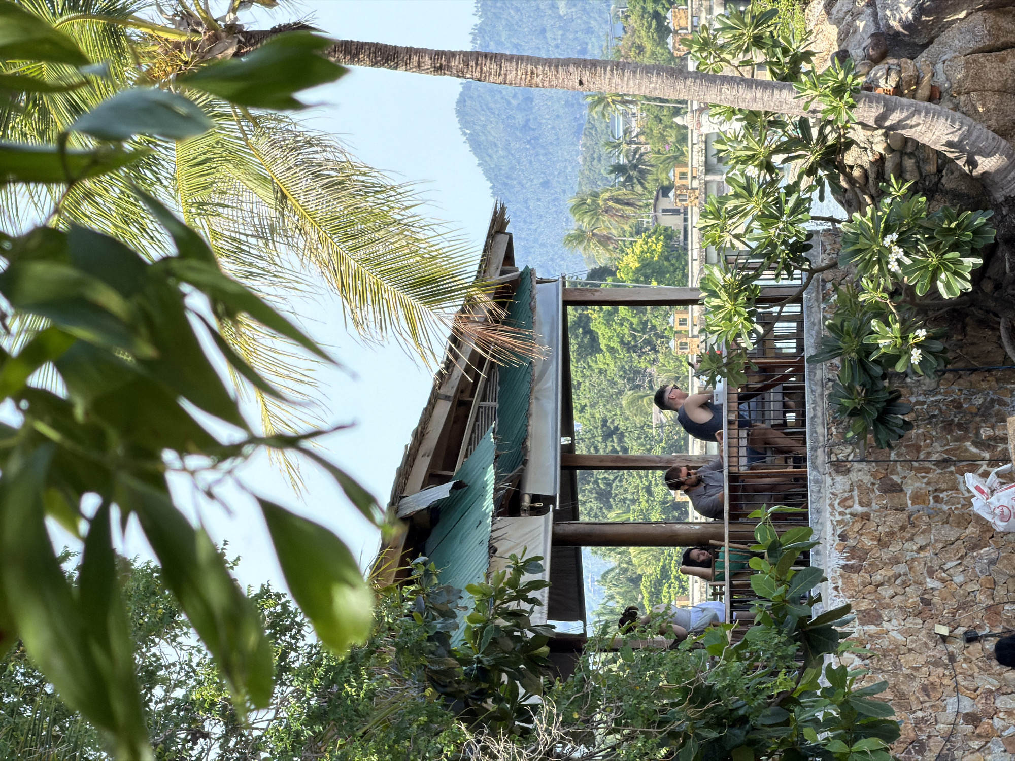 A group of people are gathered at an open-air restaurant in Baan Plai Laem, Thailand, with lush greenery and a palm tree in the foreground. The restaurant has a green metal roof and wooden pillars, with tables and chairs visible on a raised platform. People are seated at the tables, some appearing to be in conversation. Beyond the restaurant, a body of water is visible, with a bridge and buildings in the distance, and rolling hills covered in trees forming the background under a clear blue sky. The scene is framed by the leaves of trees and a large palm tree trunk.