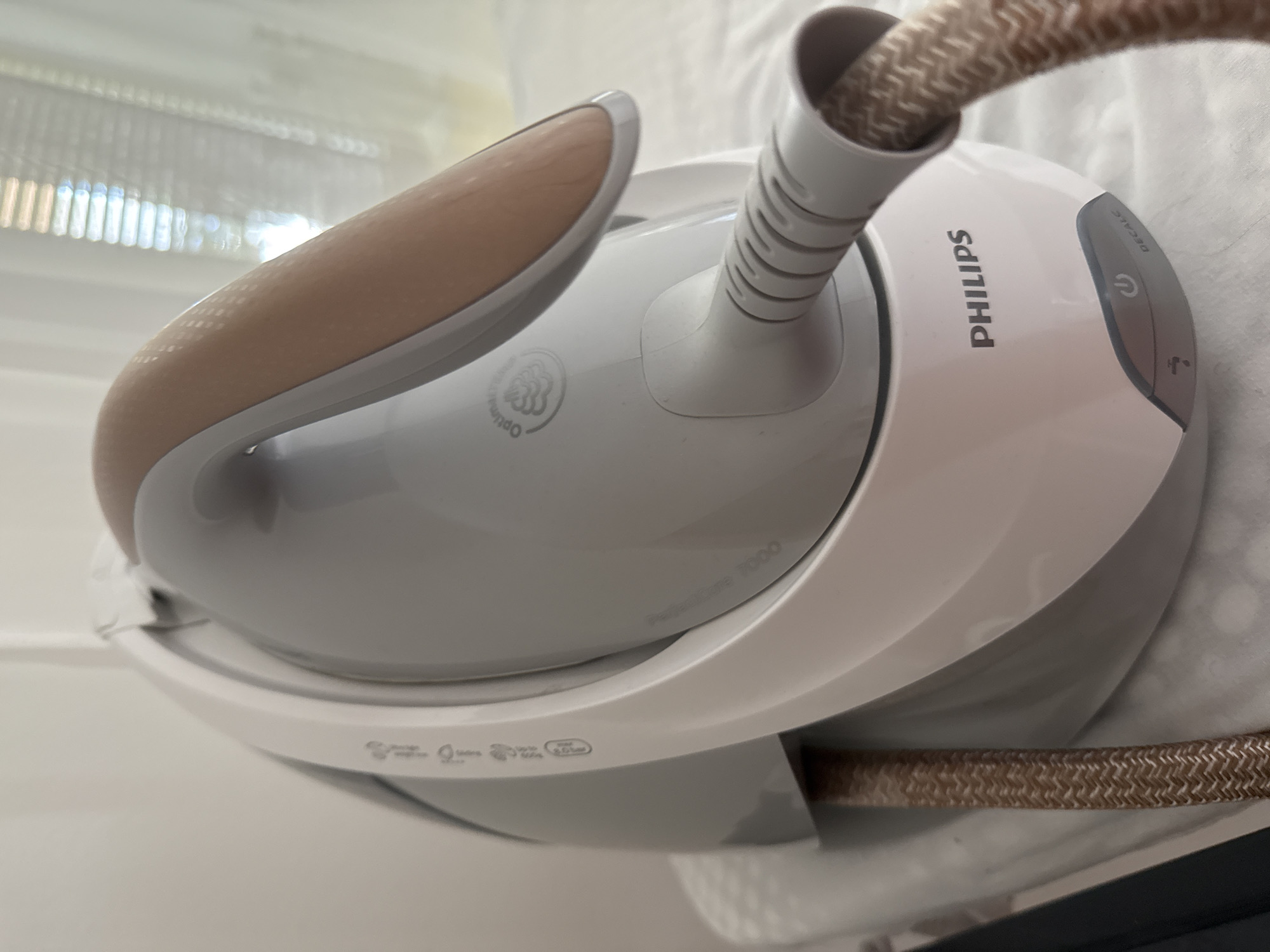 The image shows a close-up, eye-level view of a white and gray Philips steam iron with a rose-gold colored handle. The iron is situated on what appears to be an ironing board or a white, textured surface, with a white sheer curtain and blinds visible in the blurred background, suggesting an indoor setting. The iron has the brand name "PHILIPS" printed on its surface, along with icons indicating power and a descaling function. A textured, brown and white braided cord extends from the iron. No people or actions are depicted. The location is not specified within the image itself, so its connection to Găvănoasa, Moldova is not evident from the visual information provided.