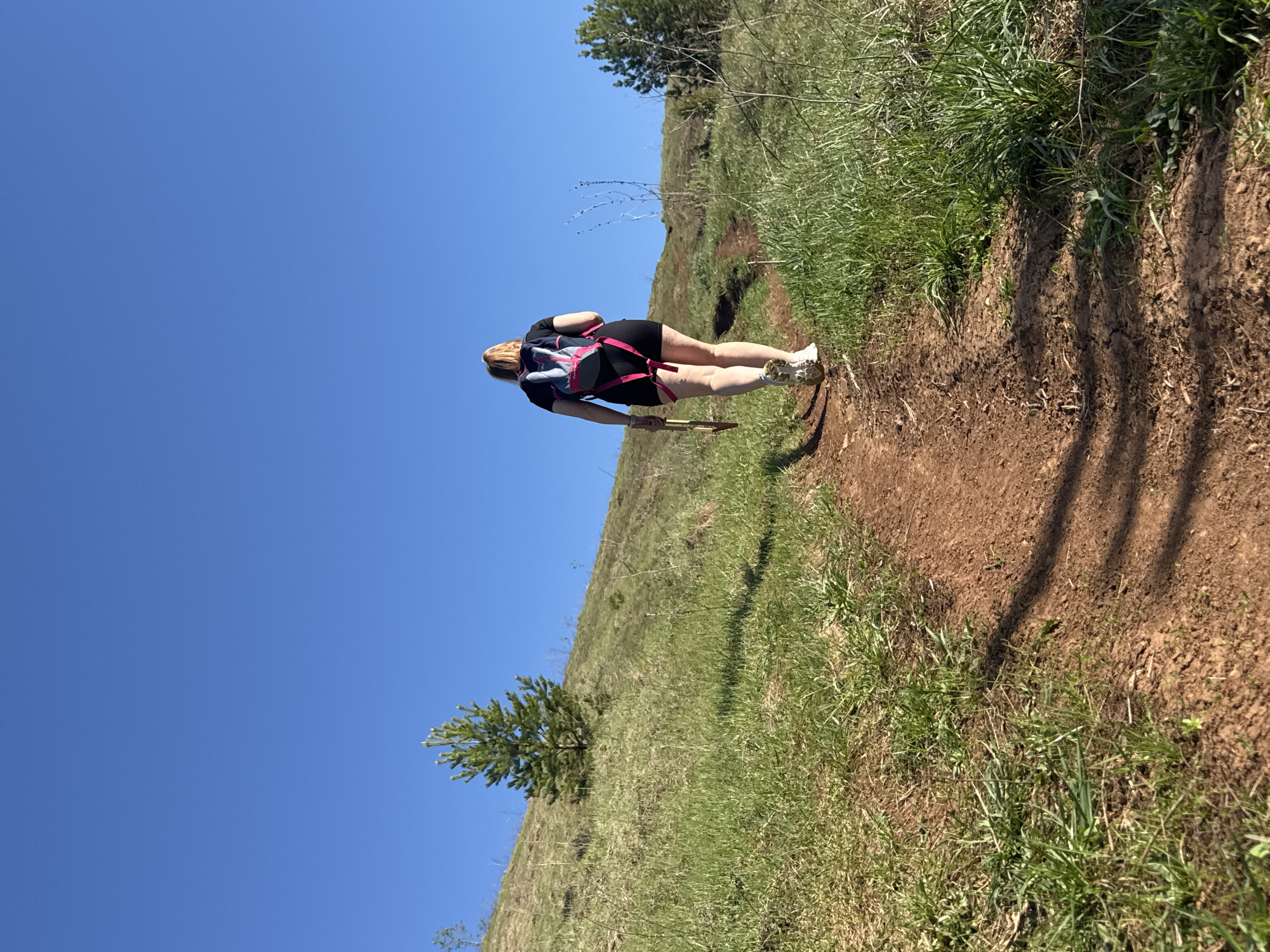 A medium, eye-level shot depicts a woman hiking on a dirt trail in Ольховка, Russia. She is seen from the back, facing slightly away from the viewer. She wears a black shirt, black shorts, and white sneakers. A pink and blue backpack is strapped to her back, and she holds a wooden stick in her right hand. The trail is bordered by green grass and a few small trees. The sky is clear and blue. The picture appears to be taken during daylight hours.