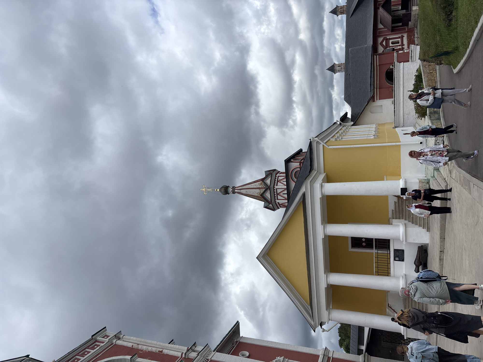 The image captures a frontal view of a yellow building with white columns and a staircase leading to its entrance. To the left, a red building with white architectural details is partially visible. In the background, a red building with a dark roof and a distinctive dome structure topped with a golden cross is prominent. The sky is overcast with heavy, dark clouds.
Several individuals are present in the scene. On the steps of the yellow building, a group of people are seen, some facing away from the camera, others conversing. To the right of the yellow building, a path curves through a grassy area, with more people walking along it.
The architecture suggests a religious or historical site. The presence of a cannon near the entrance of the yellow building implies it might be part of a museum or historical complex. The overall atmosphere is one of a cloudy day at a significant location. This scene is consistent with the Savvino-Storozhevsky Monastery in Zvenigorod, Russia, a well-known monastic complex with a rich history.