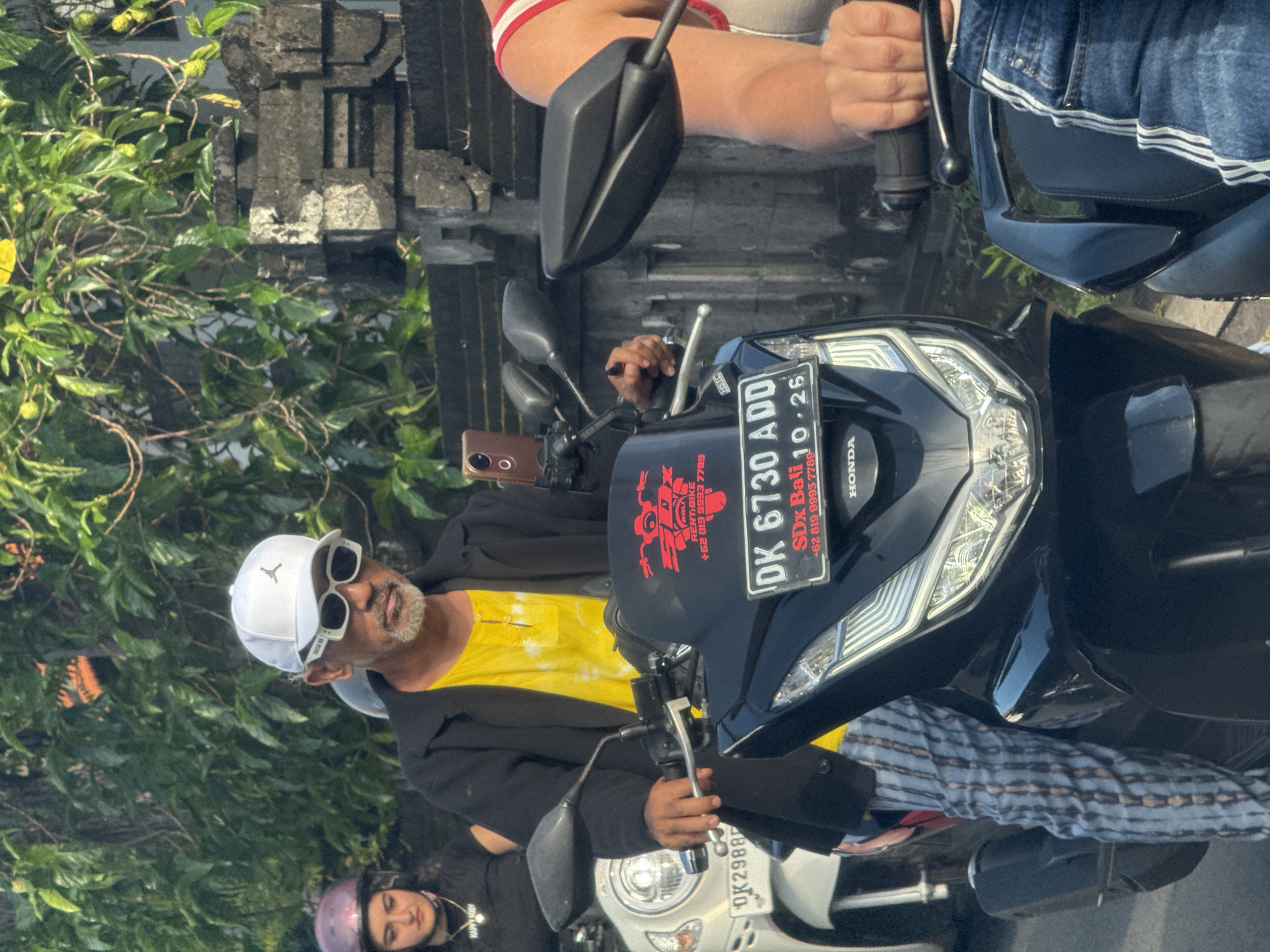The image captures a scene in North Kuta, Indonesia. In the foreground, a middle-aged man wearing a white baseball cap, sunglasses, a yellow t-shirt, and a black jacket is sitting on a black Honda scooter. The license plate of the scooter reads "DK 6730 ADD." A red and black sticker on the scooter advertises "SDx Bali RENTIBIKE +62 819 9993 7789." To the right of the man on the scooter is another person sitting on a scooter. A woman wearing a helmet and a black shirt can be seen on a scooter behind the first scooter. The environment includes greenery and a gray concrete structure, suggestive of a street setting in North Kuta. The scooters appear to be parked or stopped on the side of the road.