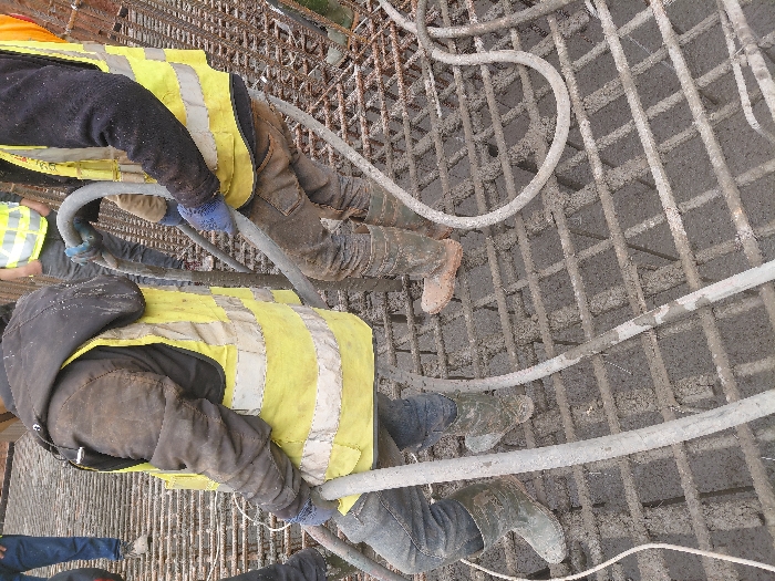 The image captures a construction scene depicting a concrete pouring operation. While the specific location within Fdalate, Morocco, is not discernible, the environment is a structure composed of a steel rebar grid partially covered with freshly poured wet concrete. At least four individuals are present. In the foreground, two male construction workers are actively engaged in directing thick, grey concrete pump hoses. Both workers wear yellow high-visibility safety vests over long-sleeved workwear; one wears a brown jacket with a black hood, and the other wears a black sweater. They are dressed in dark work trousers and protective rubber boots, which are soiled with concrete and mud. One worker wears blue gloves. The hoses are dispensing grey, wet concrete onto the rebar grid, which serves as reinforcement for the structure being formed. Other workers are partially visible in the background, also wearing high-visibility vests.