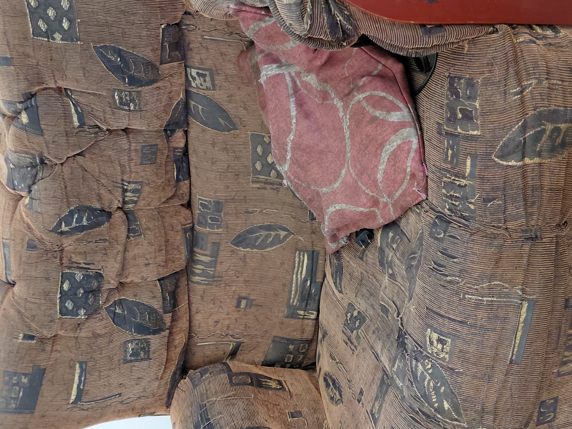 This image depicts a section of a heavily worn upholstered chair or sofa located indoors in Unknown, Unknown. The furniture is covered in a brown, vertically ridged fabric resembling corduroy, patterned with dark brown or black abstract shapes, including leaves and geometric squares, some accented with faded gold or tan details. The fabric exhibits significant wear, fraying, and flattening of the ridges across the back cushions, armrest, and seat. The back cushions appear tufted, creating folds and depressions in the material. A small, reddish-pink throw pillow with a light-colored, abstract circular pattern rests on the seat cushion, leaning against the right side. A black electrical plug or similar small dark object is partially visible next to the pillow. A portion of a solid red surface, possibly a wooden frame or an adjacent object, is visible along the far right edge of the image. A plain, light-colored wall is visible in the background on the upper left. No individuals are present.