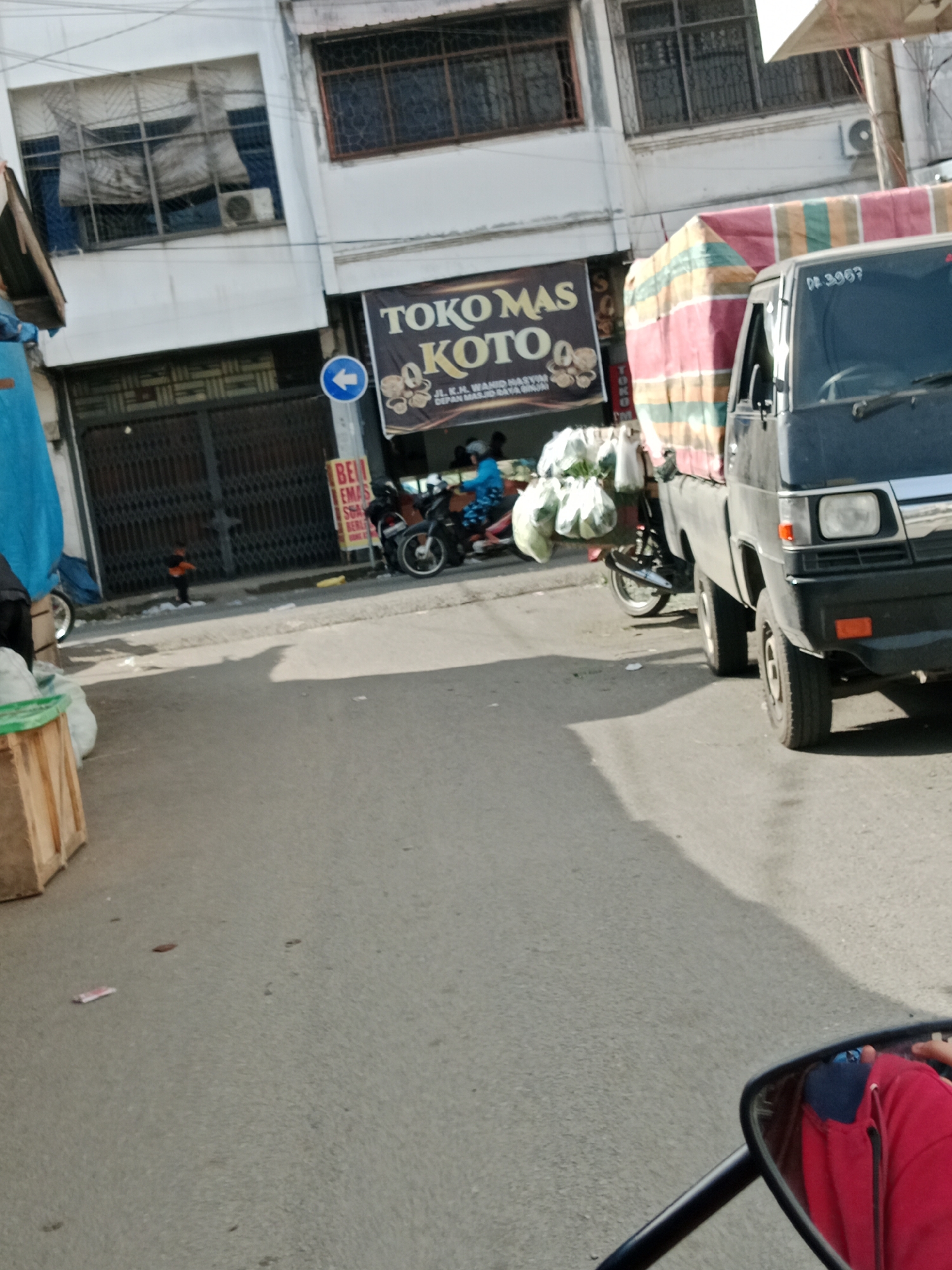 In the City of Binjai, Indonesia, a street scene unfolds with a black truck loaded with goods and a motorcycle. The truck, partially visible, features a license plate that reads "DB 3067". It is covered with a colorful striped tarp. A motorcycle is parked beside the truck, carrying several bags of produce. In the background, a building with white walls and barred windows is visible. A sign with text including "TOKO MAS KOTO" and "JL. KH. WAHID HASYIM" is displayed on the building. A blue arrow sign points left. In the foreground, a wooden crate is present, and the shadow of a person on a motorcycle, possibly the photographer, is cast on the asphalt.