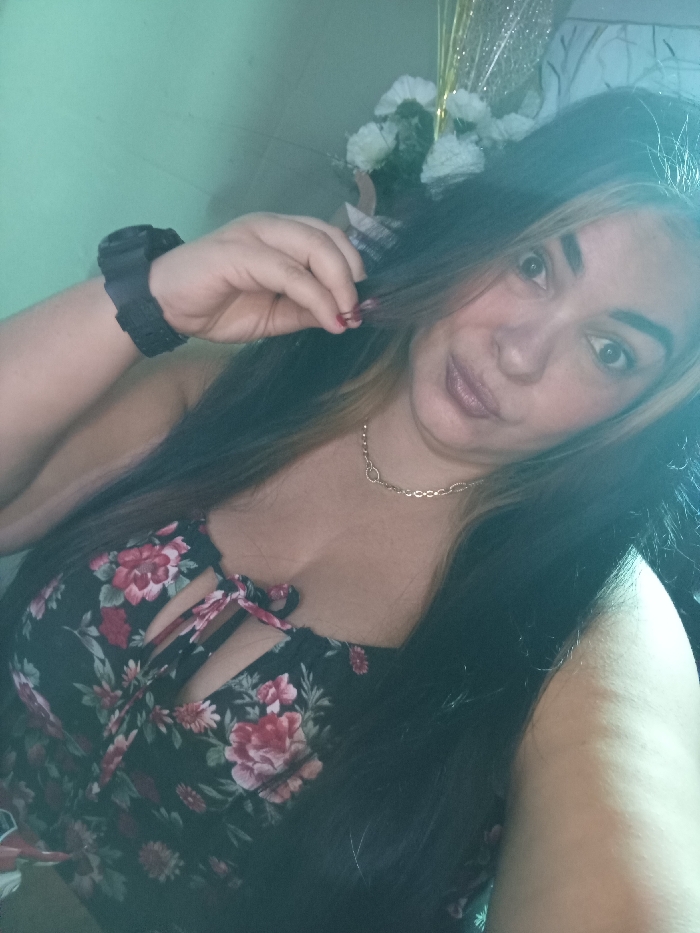 A woman takes a selfie, looking directly at the camera. She wears a floral dress and a gold necklace. She has long dark hair with a blonde streak and holds a strand of her hair with her fingers. A dark-colored scrunchie adorns her wrist. In the background, white flowers and decorative elements are visible. The lighting appears to be natural.