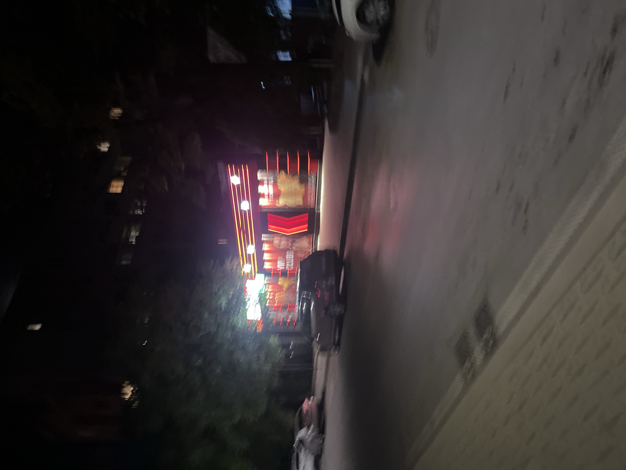 Captured at night, the image portrays a street scene in Blagoevgrad, Bulgaria. A lit-up building, presumably a nightclub or entertainment venue identified as "VIP CLUUB", dominates the view. A parked car is near the building and street lights illuminate the scene. Vegetation like trees are on the left side of the building. The street and sidewalk stretch away from the building, along the side of the road, leading towards the street corner. Buildings and trees are located on the left side of the street in the distance.
