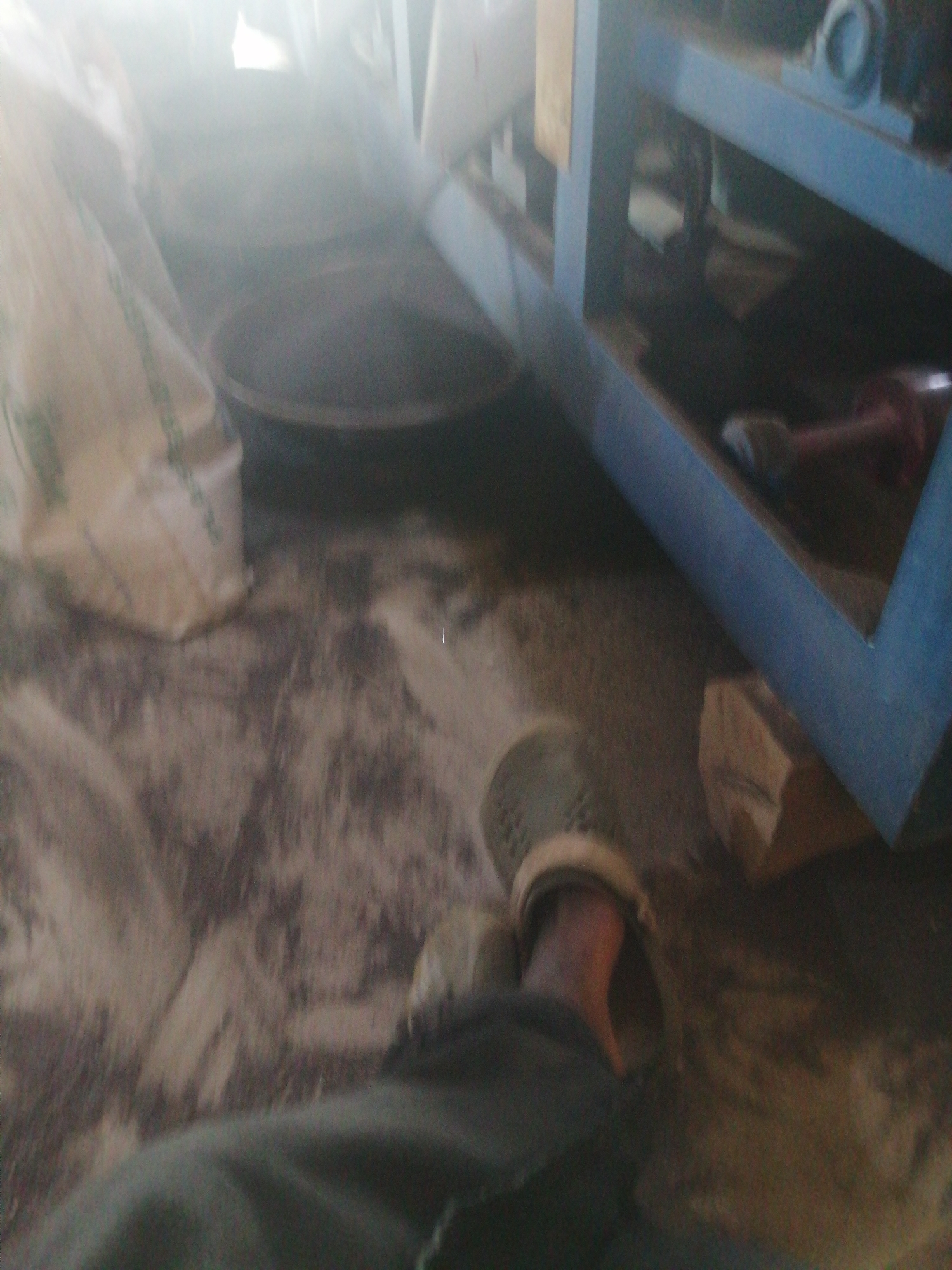 The image shows a person wearing a green garment and a light-colored shoe standing on a floor covered with sand and what appears to be scattered grains. To the left, a large, off-white bag with green stripes is visible. In the background, a blue industrial machine is present, with a red hose-like object protruding from it. The scene appears to be indoors, possibly a workshop or storage area, with dust or fine particles suspended in the air, creating a hazy effect. The lighting is bright and diffused, suggesting a strong light source from the top left. The floor is unevenly textured with a mixture of sand and lighter-colored debris. The overall impression is one of a working environment where materials like sand or grain are handled. The location is stated to be the city of Jos, Nigeria.