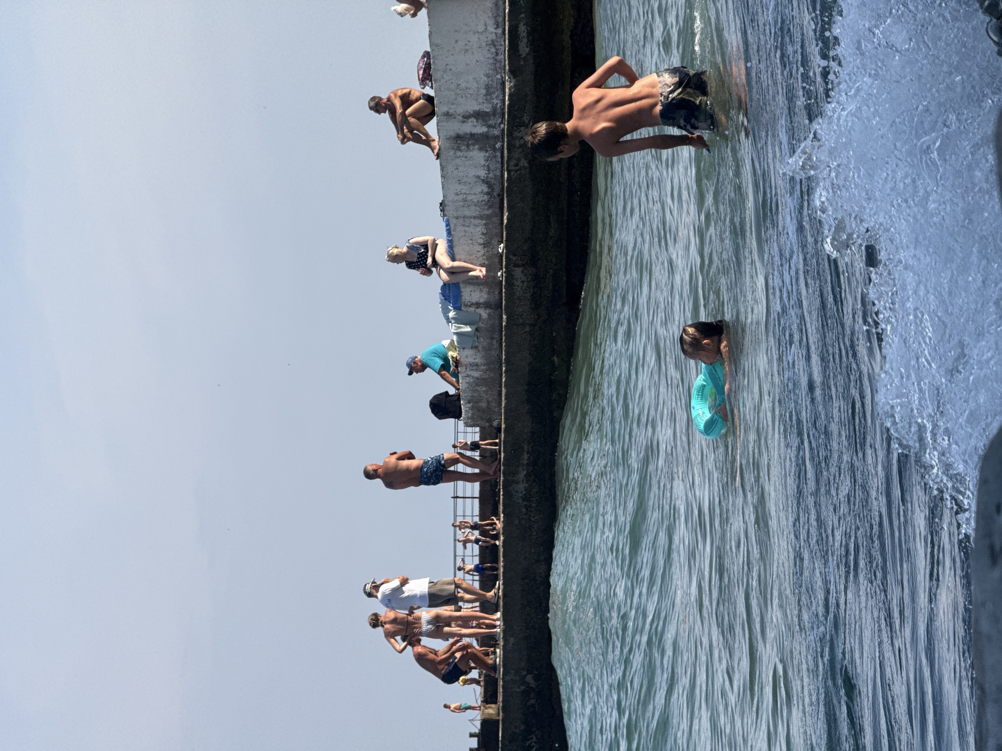 The image depicts a crowded section of a seaside promenade in Partenit, Ukraine, under a clear, bright sky. The promenade, a concrete structure, runs horizontally across the middle ground, with people scattered along its length. In the foreground, the choppy, blue-green water of the Black Sea is visible, with a wave cresting on the right.
On the promenade, various individuals are engaged in different activities. On the left, a group of people are gathered, some standing, some appearing to be in conversation. Further to the right, a man in blue shorts stands with his arms crossed. A woman with dark hair and a patterned swimsuit sits on the edge of the promenade, while a man in dark shorts and sunglasses sits nearby. Other individuals are visible further down the promenade, some standing and some seated. A backpack rests on the concrete.
In the water, two children are present. One child, wearing a blue inflatable ring, is swimming, with only their head and shoulders visible. Another child, a boy, stands in the water, facing away from the viewer. His back is to the camera, and he appears to be partially submerged. The overall scene suggests a warm, sunny day at a popular beach resort.