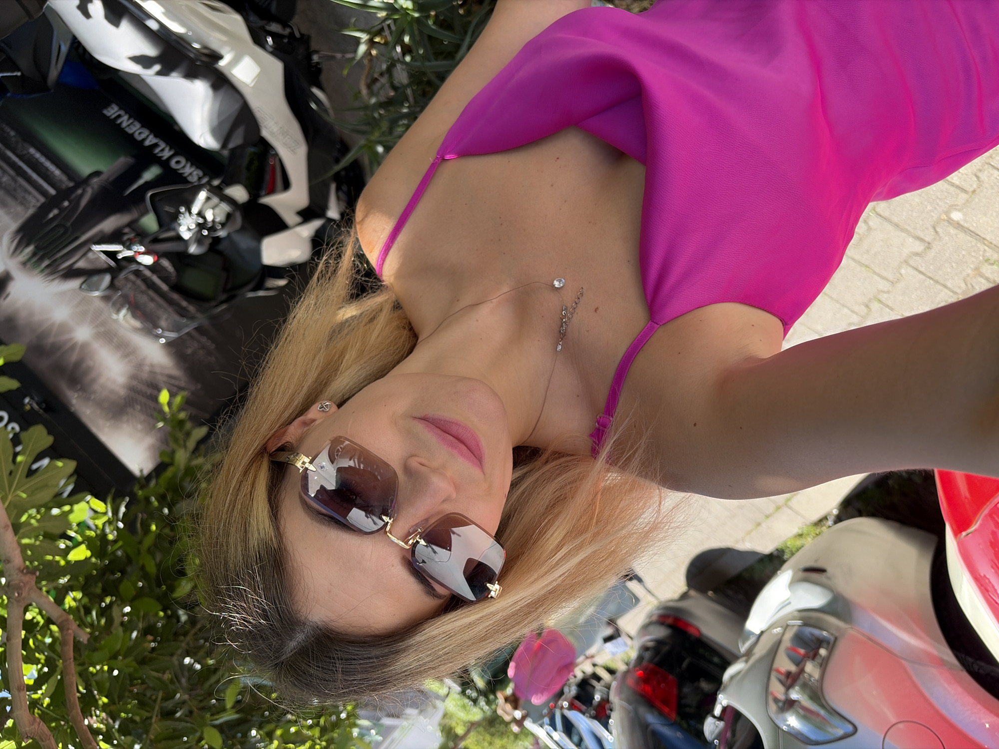 This is a slightly tilted, medium shot selfie taken outdoors on a sunny day. The subject is a woman with blonde hair and sunglasses, wearing a bright pink dress. She is looking towards the camera, and her expression is relaxed and perhaps a little playful. A delicate necklace with a small pendant is visible on her neck.
Behind her, the scene is a bit cluttered but suggests an outdoor environment. There are glimpses of a motorcycle in the background, with white and black detailing and some visible text on its side. To the right, parts of other vehicles, possibly scooters or motorcycles, are visible, one in white and another with a red section. Some greenery, likely bushes or trees, frames the left side of the image, with sunlight filtering through the leaves. The ground appears to be paved with light-colored stones, suggesting a pathway or street.
The overall impression is a casual, sunny day outing, perhaps in a tourist area given the presence of multiple vehicles and the Mediterranean-style paving. The woman's attire and sunglasses add to a summery, vacation-like vibe. The specific text visible on the motorcycle is "RIČKO KLADENJE".