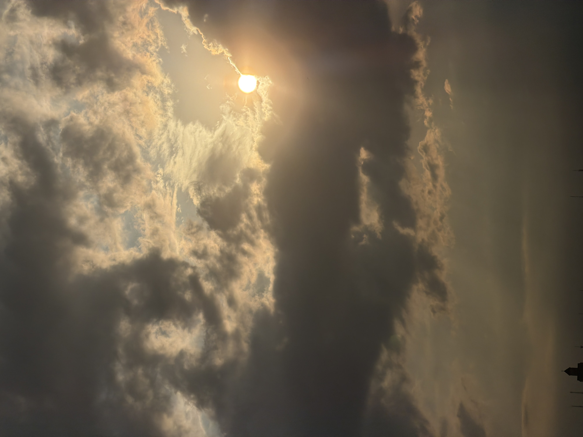 The image shows a cloudy sky with the sun peeking through the clouds. The lighting suggests either sunrise or sunset. In the lower right corner, a silhouette of buildings and structures is visible, which aligns with the architecture found in Tbilisi, Georgia. Due to the limited information, a detailed description of specific people or actions isn't possible. The main subject of the image is the dramatic sky and the interplay of sunlight and clouds, likely captured at a moment near dawn or dusk.