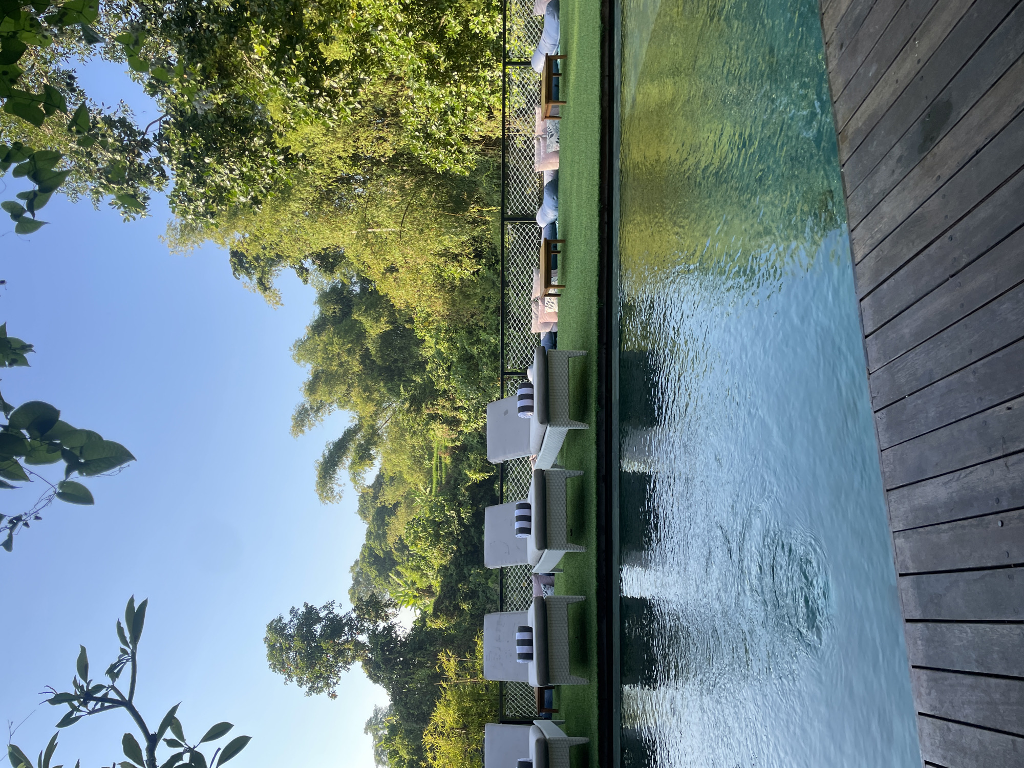 This image depicts an outdoor swimming pool area surrounded by lush greenery, suggesting a resort or private villa setting. The pool itself is in the foreground, with a wooden deck bordering it. In the background, several white lounge chairs are neatly arranged on a patch of artificial grass, each adorned with striped pillows. Beyond the lounge chairs, there's a seating area with large floor cushions and low wooden tables. A black metal fence separates this area from the dense tropical foliage, which includes bamboo and various trees. The sky is clear blue, indicating fair weather. There are no people visible in the scene. The location is described as Kediri, Indonesia.