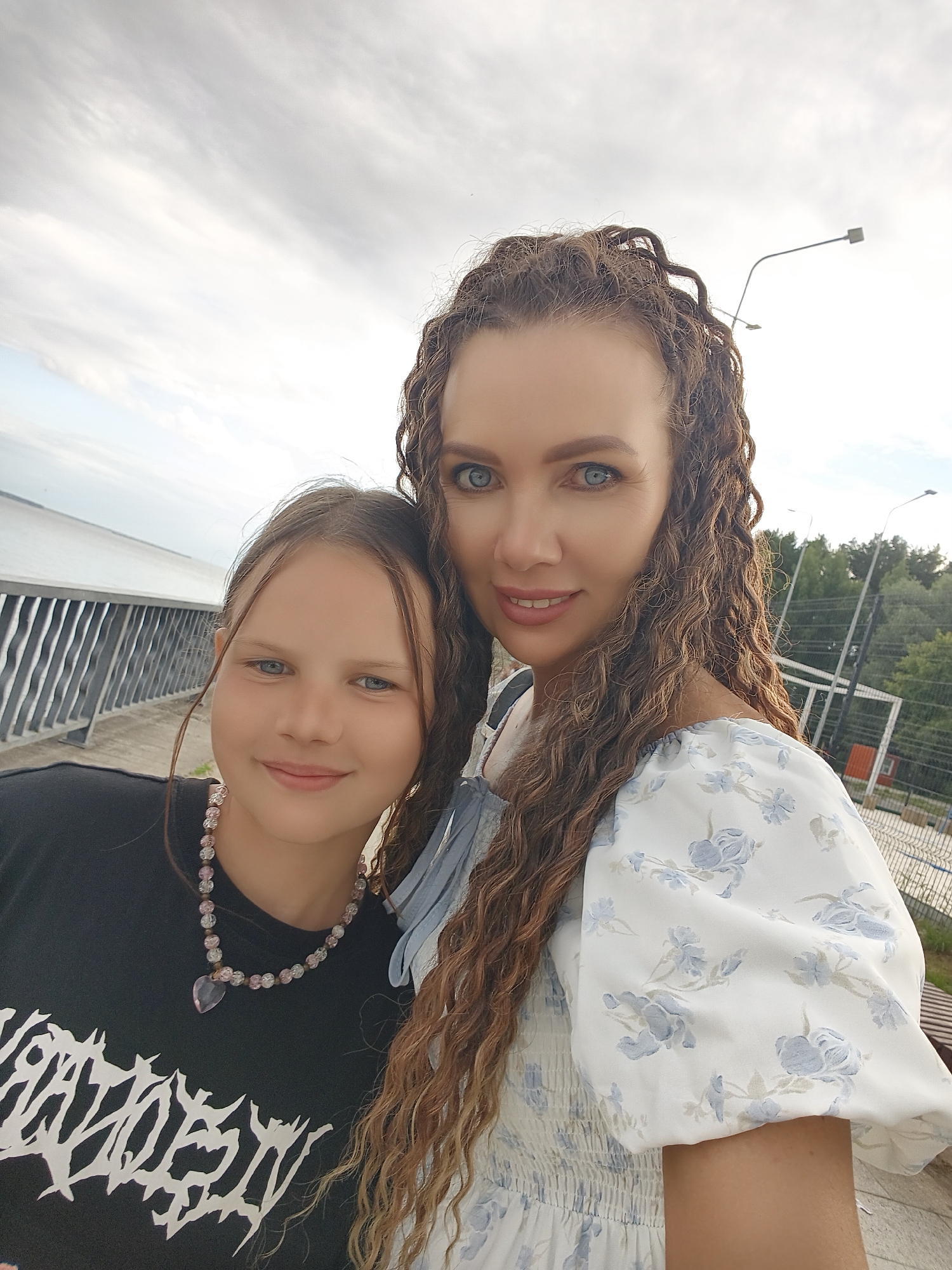 A selfie taken outdoors features two females. The person on the left is a young girl with light brown hair, blue eyes, and a light complexion. She is wearing a black t-shirt with a white graphic design and a beaded necklace with a pink heart-shaped pendant. The person on the right is a woman with long, curly brown hair, blue eyes, and a light complexion. She is wearing a white dress with a blue floral pattern and puff sleeves. They are standing on a paved area with a metal railing to their left and a fence with trees and lights in the background. The setting appears to be a public space near a body of water. This location is the embankment of the Kama River in the city of Chaikovsky, Russia.