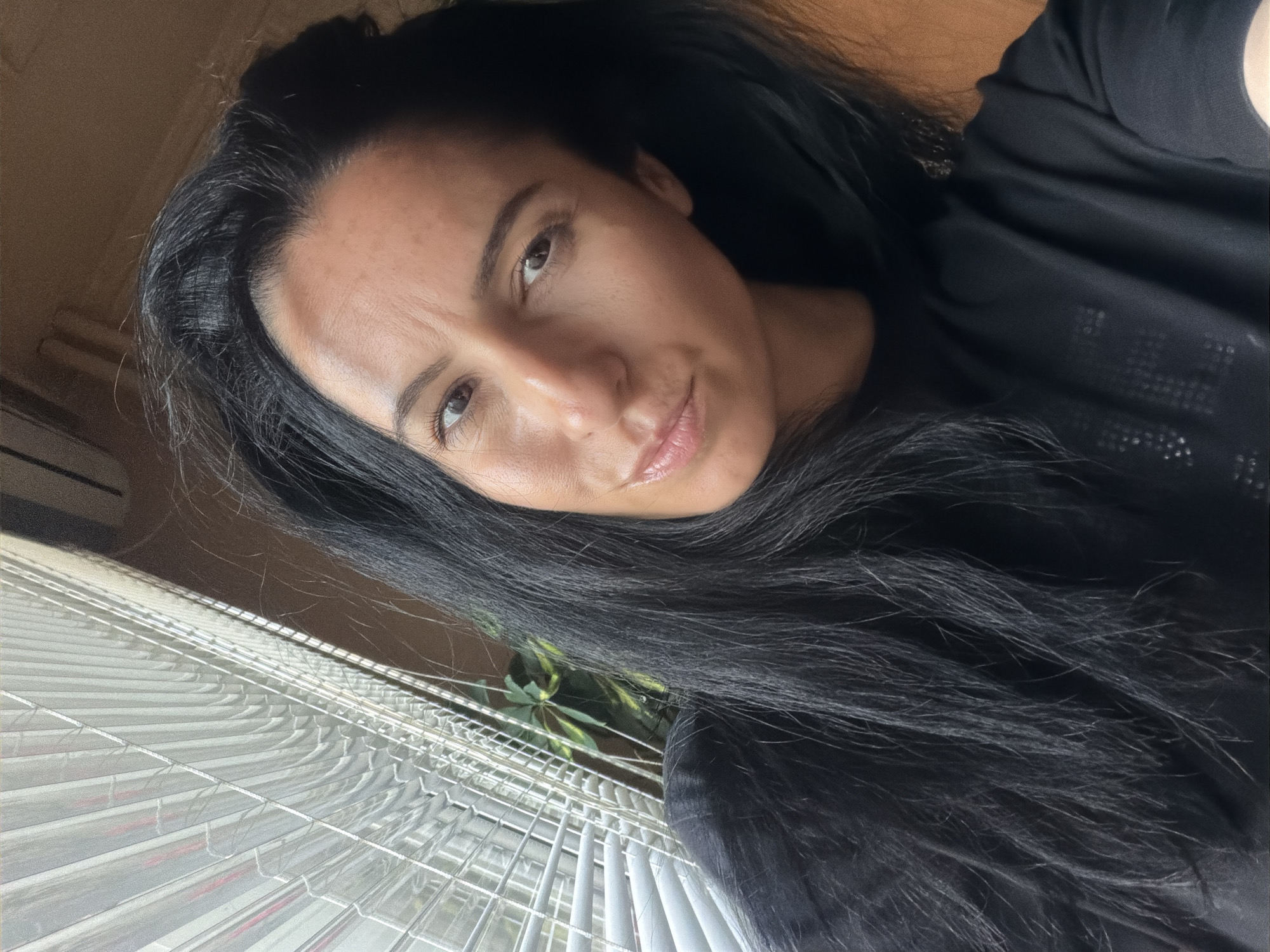 A woman with dark, long hair is the subject of the selfie. She's wearing a black shirt and positioned at an angle, looking slightly upward towards the camera. The background includes a window with white blinds. Partially visible behind the blinds is a green plant. The location and city of Pleven, Bulgaria, are not discernible from the image.