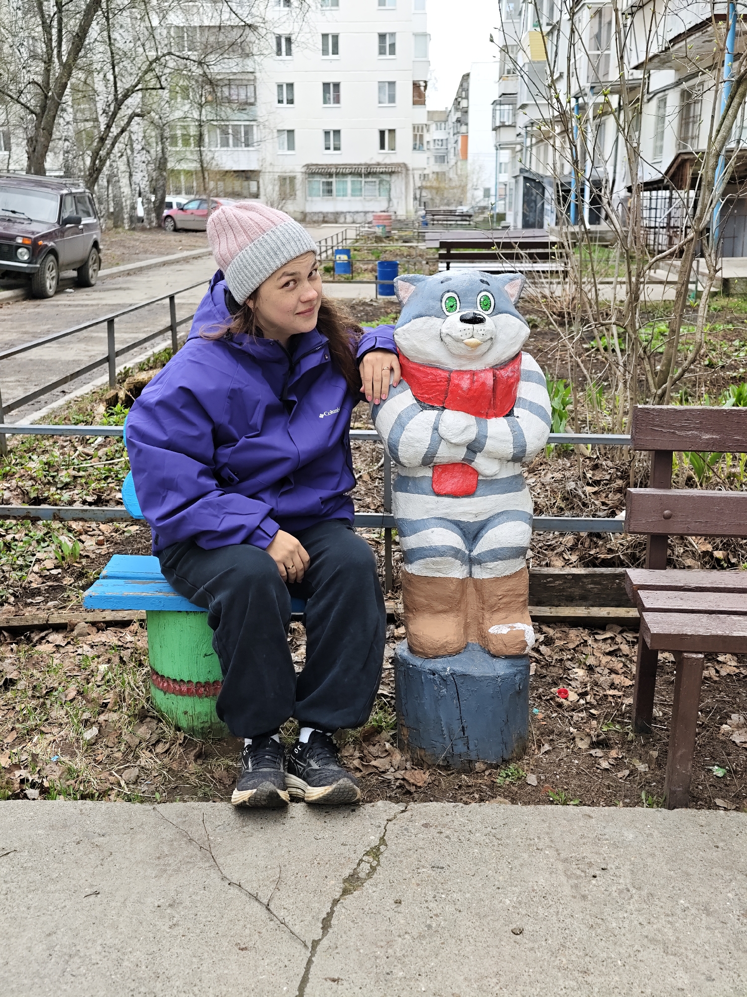A woman is seated on a small, brightly painted blue and green bench, posing next to a whimsical cat statue in what appears to be a residential courtyard. The woman, with brown hair and fair skin, is looking towards the camera with a slight smile. She is wearing a dark purple insulated jacket with a "Columbia" logo on the left sleeve, black pants, a pink and grey knitted beanie, and black sneakers. Her right arm rests gently on the shoulder of the statue.
The statue depicts a smiling grey and white striped cat with large green eyes and a bright red scarf around its neck and waist. It stands on a blue-painted tree stump and has brown, boot-like feet. To the right of the statue is another rustic brown wooden bench. The ground in the foreground consists of a concrete path, followed by dirt and a scattering of dry, fallen leaves with sparse green growth, indicating late winter or early spring.
In the background, tall, light-colored apartment buildings with many windows rise under an overcast sky. Bare deciduous trees are scattered throughout the scene. On the left side of the image, a dark-colored SUV is parked near a metal fence, with other vehicles visible further back. The scene has a typical urban residential feel, with diffused natural light suggesting it is daytime. No other significant text is visible.