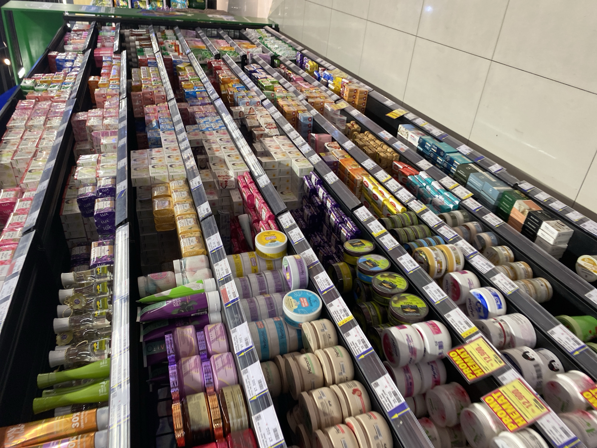 The image shows a well-stocked supermarket aisle in Banda Aceh, Indonesia, with shelves filled with various cosmetic and personal care products. The products are arranged in a neat and organized manner, with most of them packaged in boxes and bottles. The shelves are made of dark metal and are illuminated by fluorescent lights, creating a bright and clean environment. Some of the products visible include lotions, creams, soaps, and other beauty items, with prominent labels and branding. There are price tags and promotional signs displayed on some of the shelves. The floor is tiled with large light-colored tiles, and the overall scene suggests a typical retail setting. No people are visible in the image.